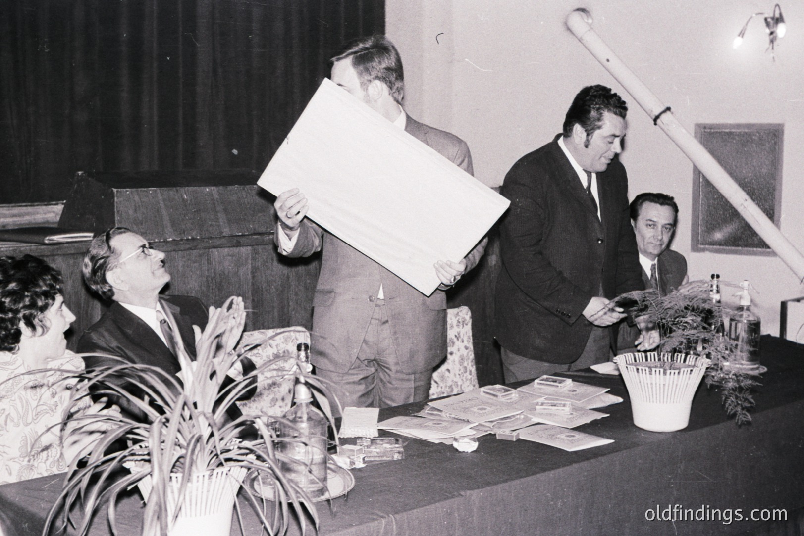 Formal meeting or presentation scene; three men and a woman seated behind a table. A large chart or display board is being presented. Papers and a plant adorn the table. Likely a 1960s-1970s setting. Potential for historical or architectural research.