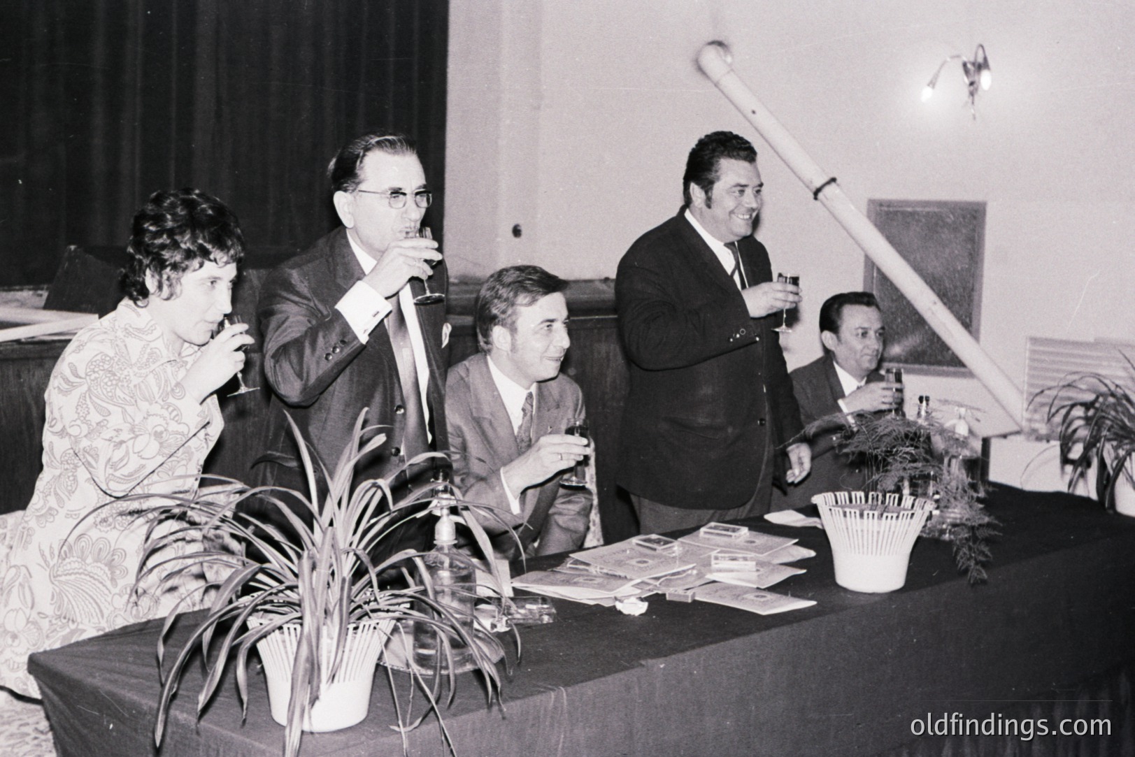Formal event scene, c. 1960s-70s. Four individuals at a table, likely judging a competition—note cards visible. Man in glasses holds drink, another smiles broadly. Décor includes potted plants and a distinctive, tall, cylindrical architectural feature.