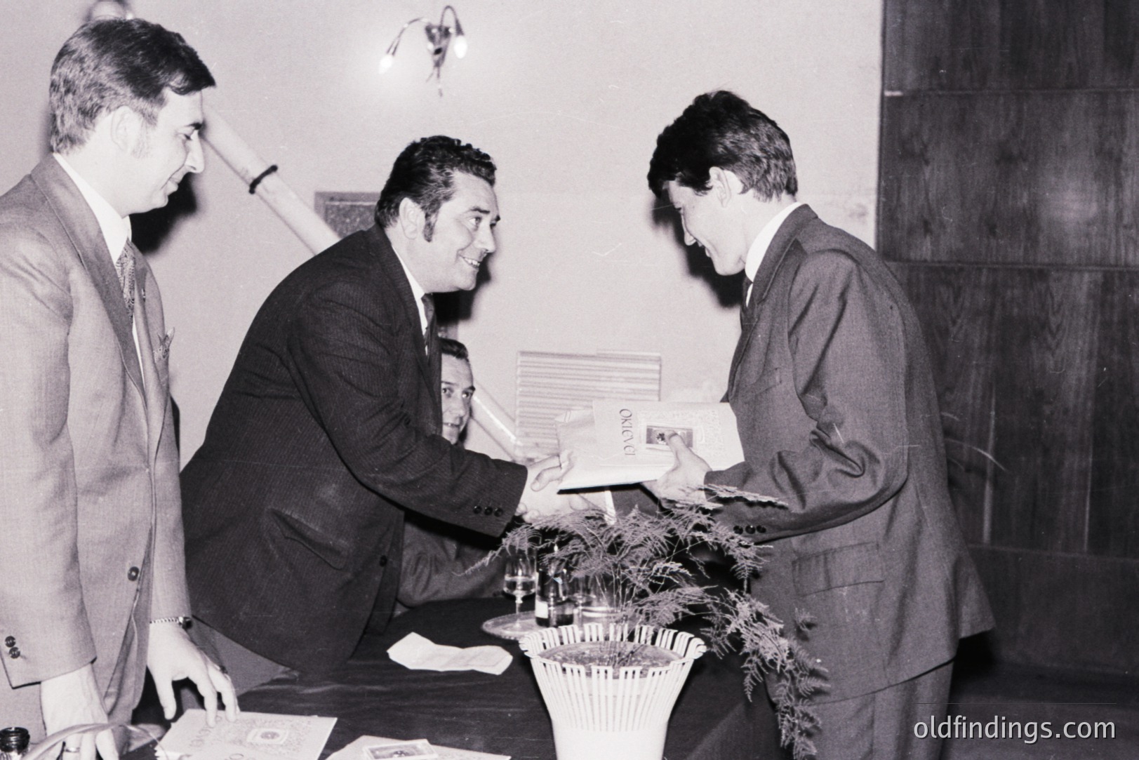 Three formally-dressed men appear during an exchange, likely a presentation or official meeting. The man in the center smiles as he hands a document to the man on the right. A plant arrangement sits on a table between them. Likely 1960s-1970s business setting.