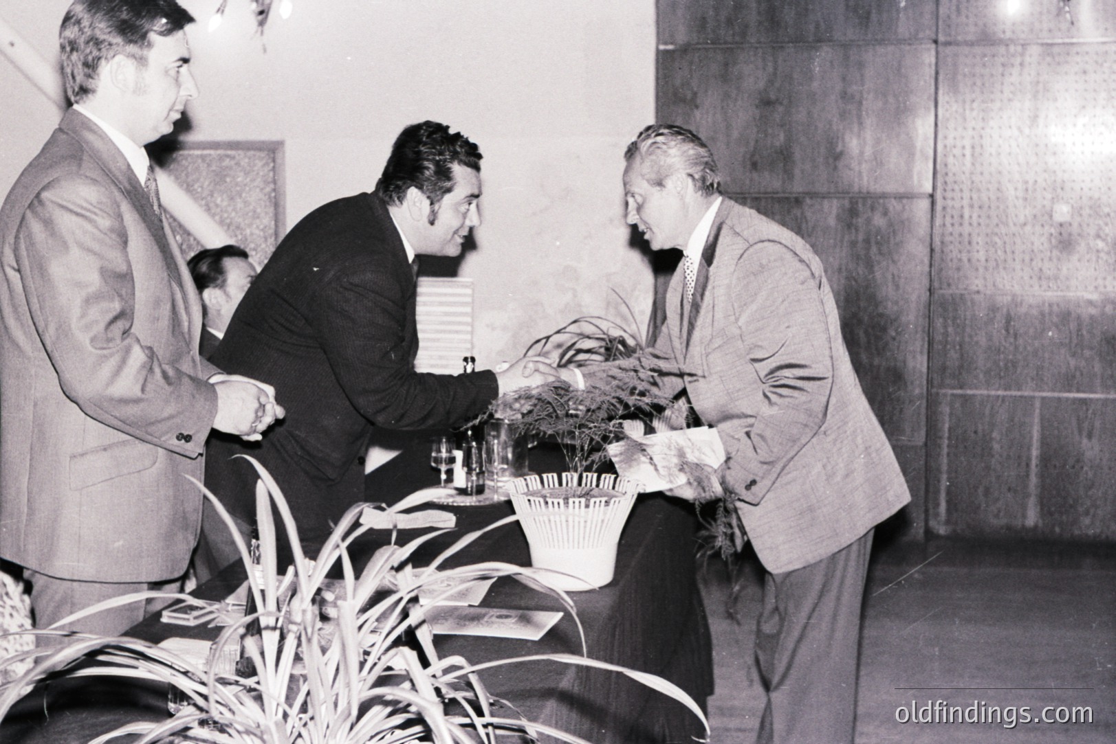 Three men in suits interact near a table covered with brochures and a decorative plant arrangement. A formal reception or awards ceremony, possibly mid-century (1960s-1970s), based on attire & photographic style. Interior space with wood paneling. Location and event details unclear. Appears archival or documentary in nature.