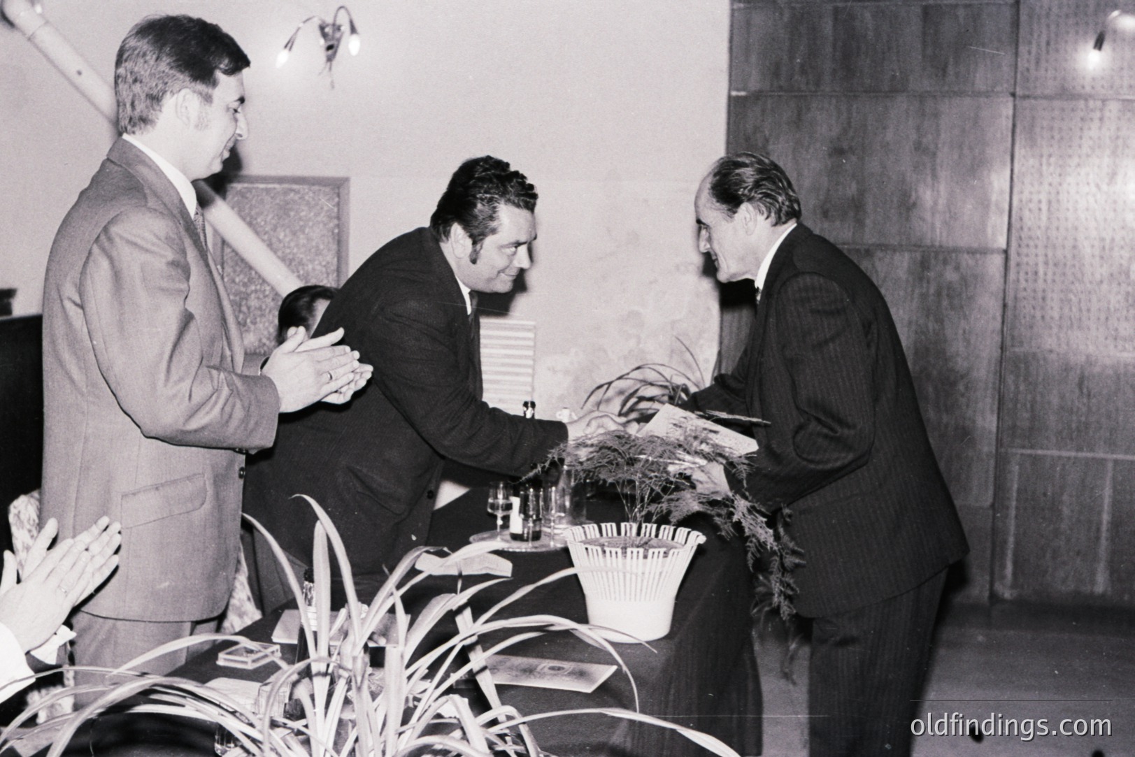 Three formally dressed men are engaged in a cordial exchange. One presents a floral arrangement over a draped table. The setting appears to be an interior space with textured wall paneling. Likely a reception or formal event. Appears to be from the 1970s.