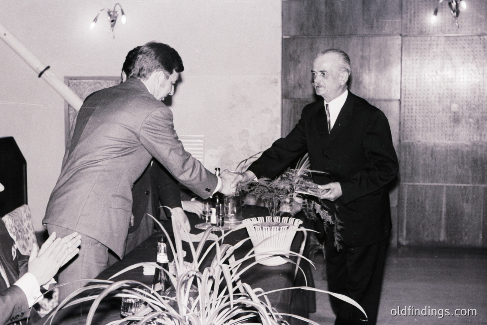 Two formally dressed men shake hands across a table adorned with foliage and glassware. Interior scene with dark wood paneling suggests a business meeting or ceremonial event. Likely 1960s or 70s, based on attire & photographic style. Potential archival value for business/social history.