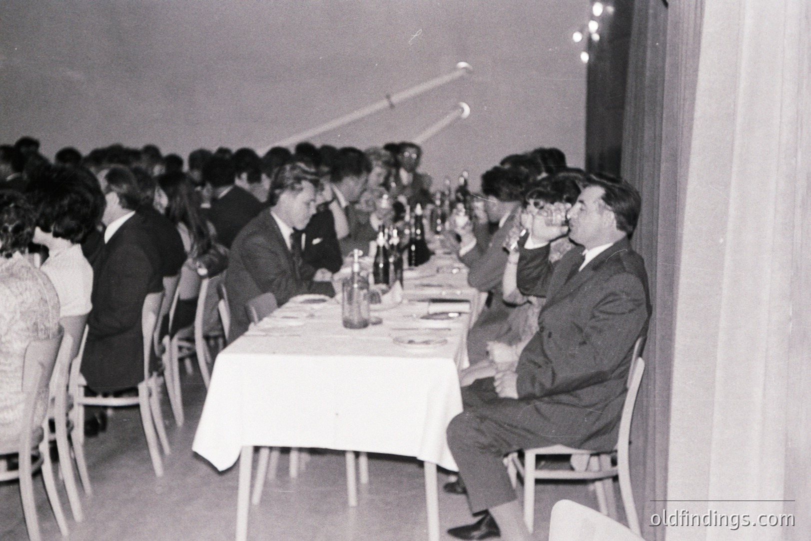Formal dinner scene, likely a banquet or reception. Guests in suits and dresses seated around a long table covered with a white tablecloth, with bottles of red wine visible. A man raises a toast, possibly champagne.