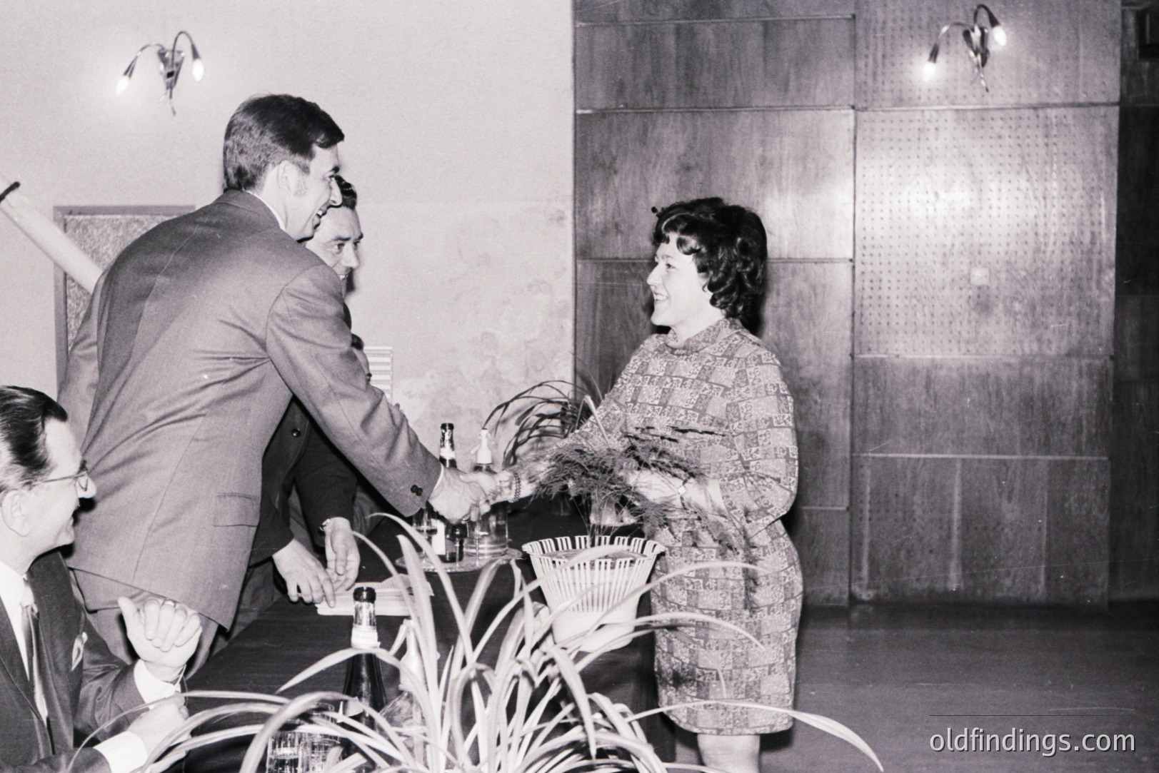 A man in a suit serves a drink to a woman in a patterned dress, likely at a social event. Interior setting features wood-paneled walls and decorative sconces, suggesting a mid-century aesthetic. A partially visible guest stands nearby. Possibly a promotional or candid shot.