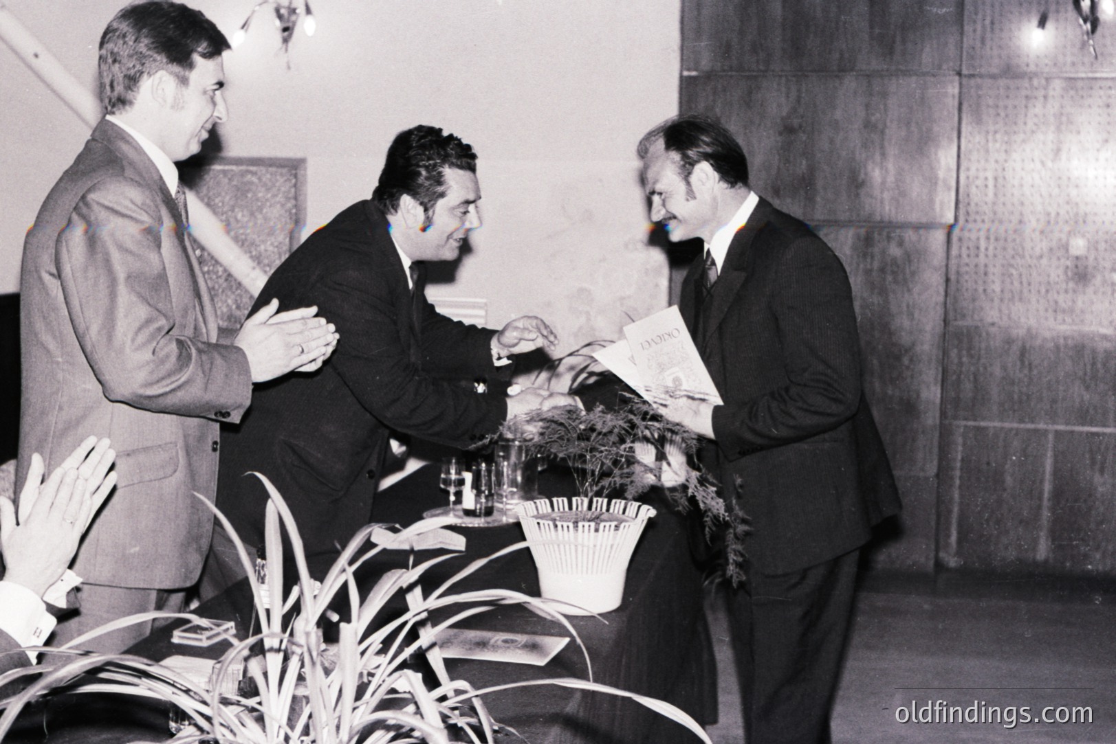 A formal presentation; two men in suits offer what appears to be a ceremonial plaque or diploma to a third, who smiles and gestures. The setting is a modern, possibly corporate, interior with a textured wall. Likely 1970s style, capturing a moment of professional recognition.