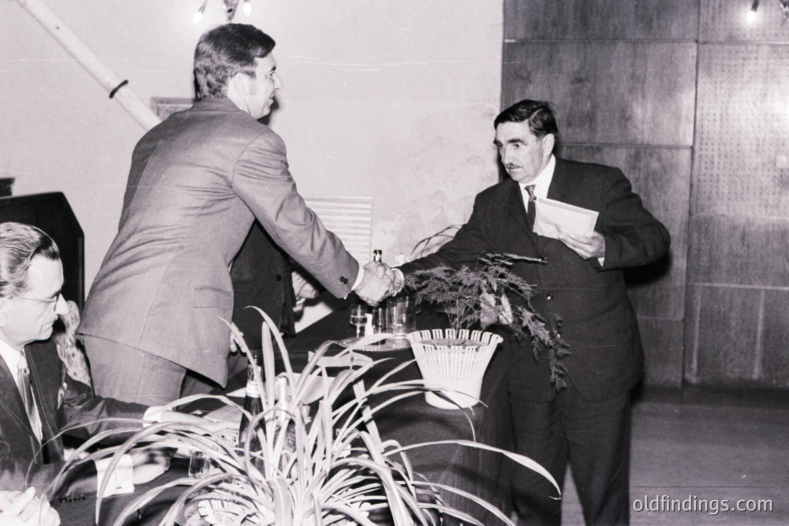 Two men share a formal handshake, one extending a document. The man on the right wears a dark suit and tie, while the other has a lighter overcoat. A blurred figure and potted plants are visible in the background. Likely a business or ceremonial event.