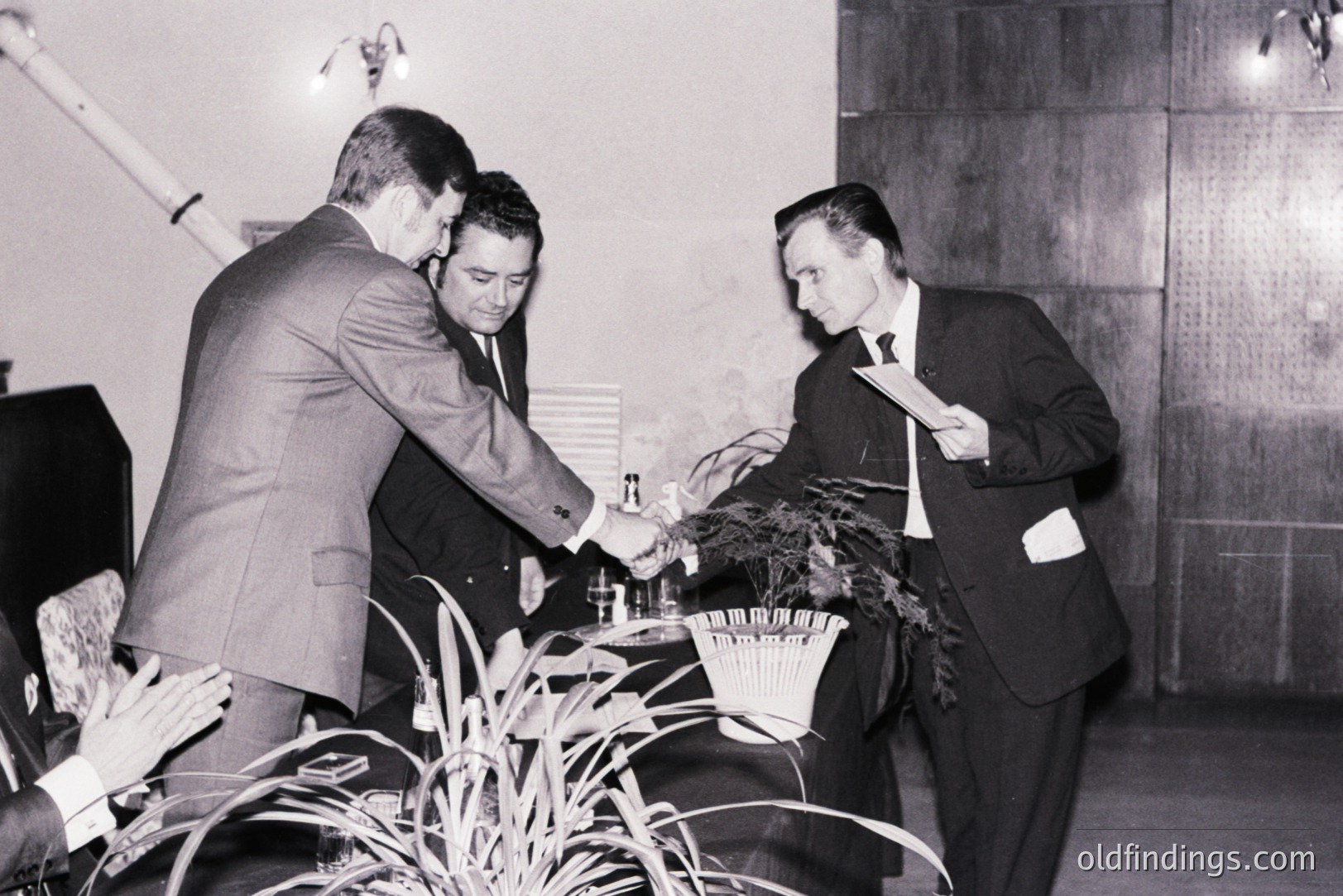 Three men in dark suits and slicked-back hair stand around a table covered with small bottles and a plant. Likely a formal event or presentation, possibly a product launch or conference. Architectural details suggest mid-century modern design.