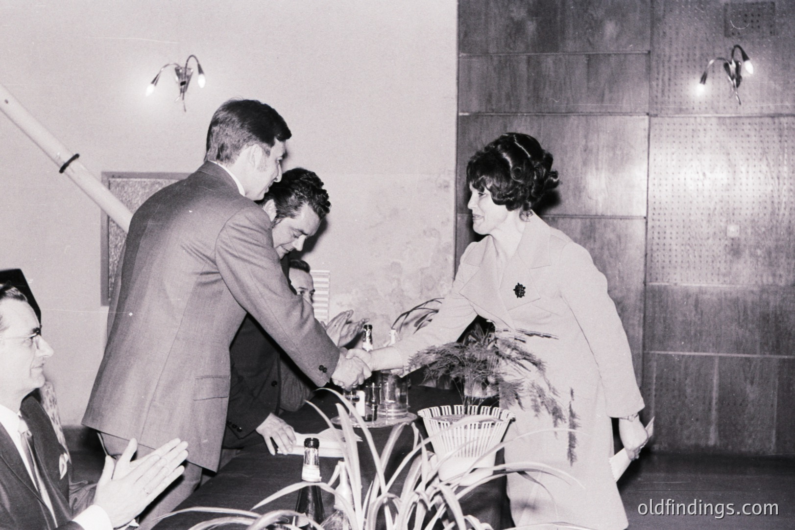 A formal presentation scene: a man in a suit receives a plaque from a woman in a tailored suit, likely at an awards ceremony or recognition event. Foliage decorates the table. A man watches from the left. Interior, likely 1960s-70s. Minimalist design. Architectural details visible.