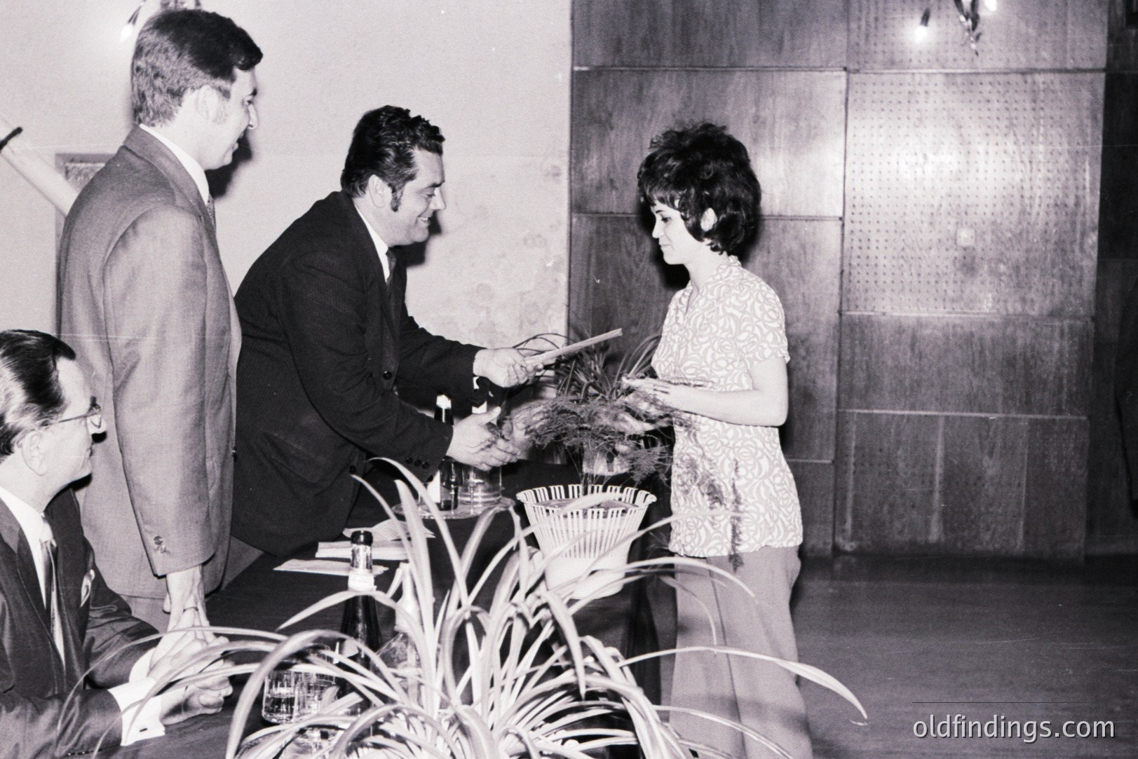 Formal portrait captures an award ceremony, likely in the 1960s. A man in a suit presents a plaque to a woman with a stylish bob haircut. Several men in suits observe the moment. The backdrop features a paneled wall and decorative plants. Classic mid-century aesthetic.