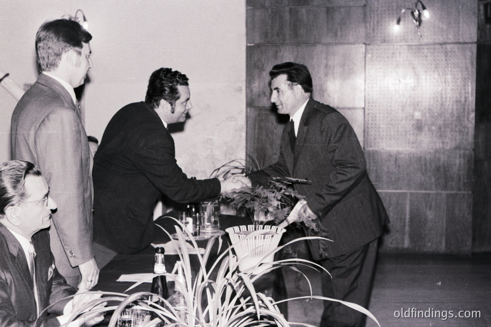 A formal meeting scene; two men in dark suits shake hands across a table adorned with greenery and a bottle of liquor. A third man observes. The backdrop features a paneled wall and sconce lighting, typical of mid-century design. Likely 1960s, possibly corporate or diplomatic setting.