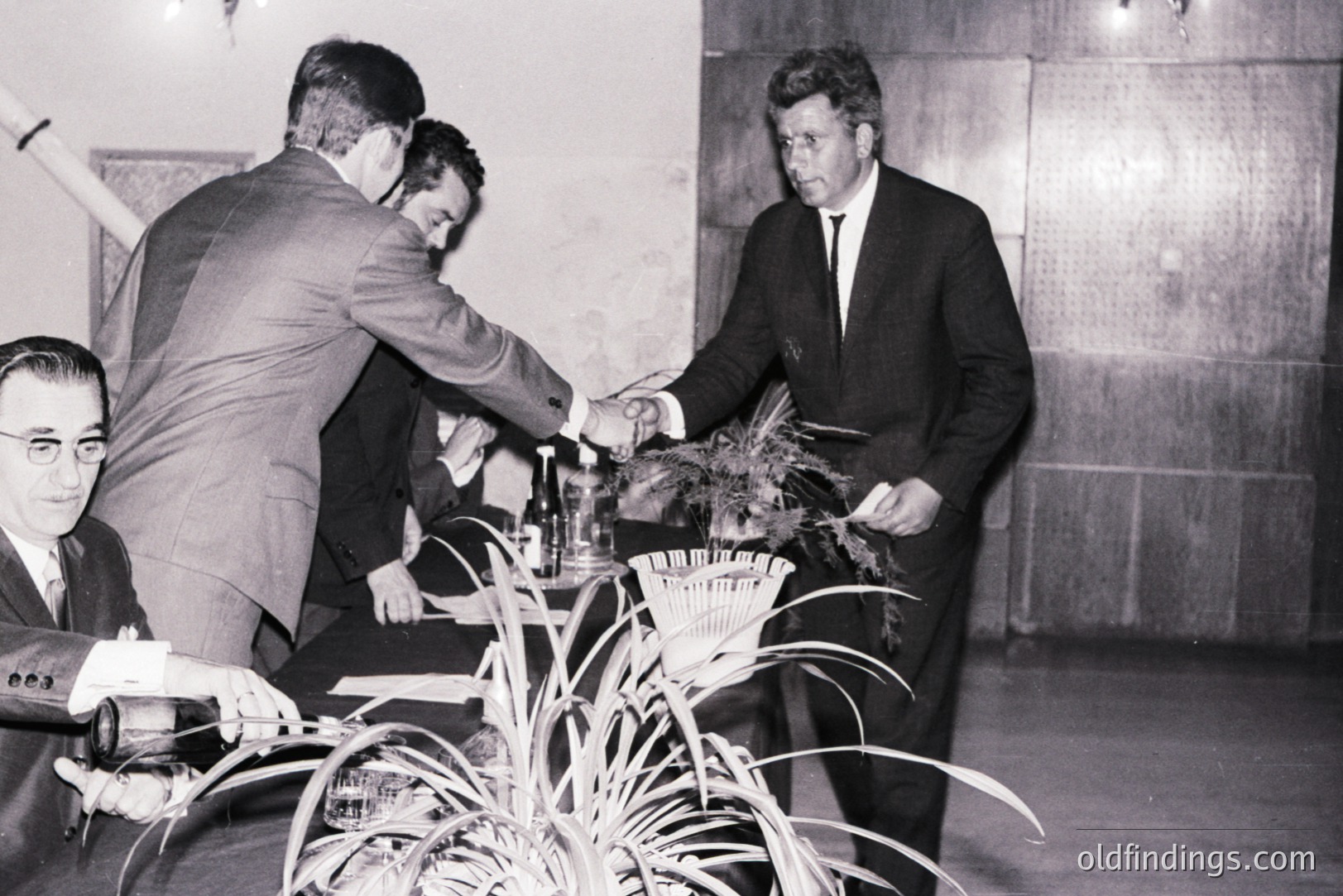 President John F. Kennedy participates in a ceremonial olive tree planting, likely symbolizing peace and prosperity. Visible are two men assisting him. A man in glasses observes. Appears to be an indoor setting with textured wall paneling. Likely mid-1960s. Valuable for historical and archival reference.