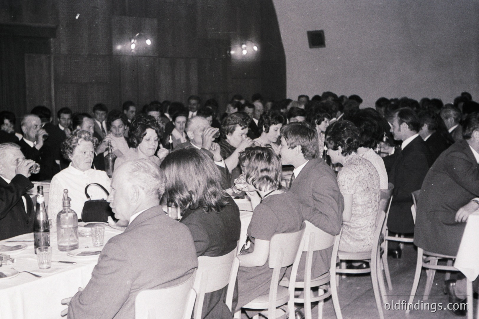 A large crowd gathered at a formal event, likely a banquet or conference. Guests are seated at tables, dressed in 1970s attire – suits, dresses, and formal wear. Noticeable darkroom equipment in background. Captures social scene & fashion.