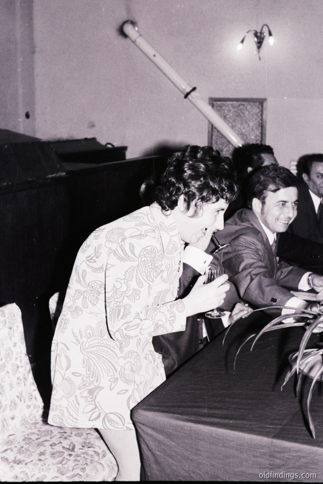 A woman in a floral print dress leans over a table, appearing to examine a document. A man in a suit smiles nearby. Likely a conference or formal event. Estimated 1960s-70s, possibly European setting. Potential use for period design or historical research.