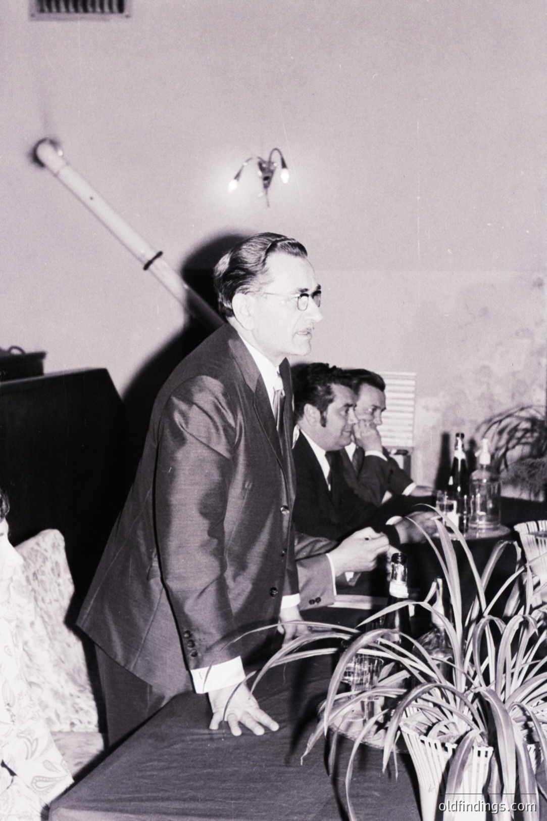 Man in a suit stands, appearing to address a seated group. A long, white object (possibly an instrument) dominates the left foreground. Tables set for a formal gathering are visible in background. Likely a staged event.