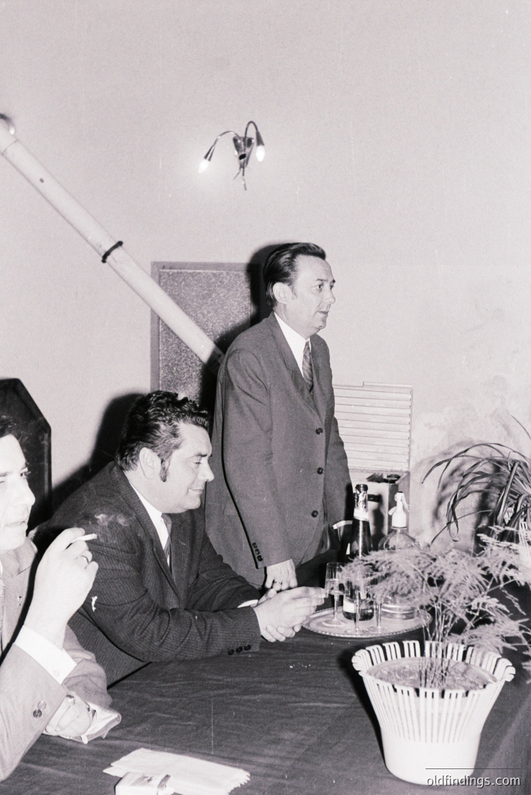 Men seated around a dark, formal table during an event. One man is smoking, another is speaking while gesturing. A potted palm adds greenery. Likely a business meeting or formal gathering, 1950s-1970s style. Architectural details include paneling & lighting.