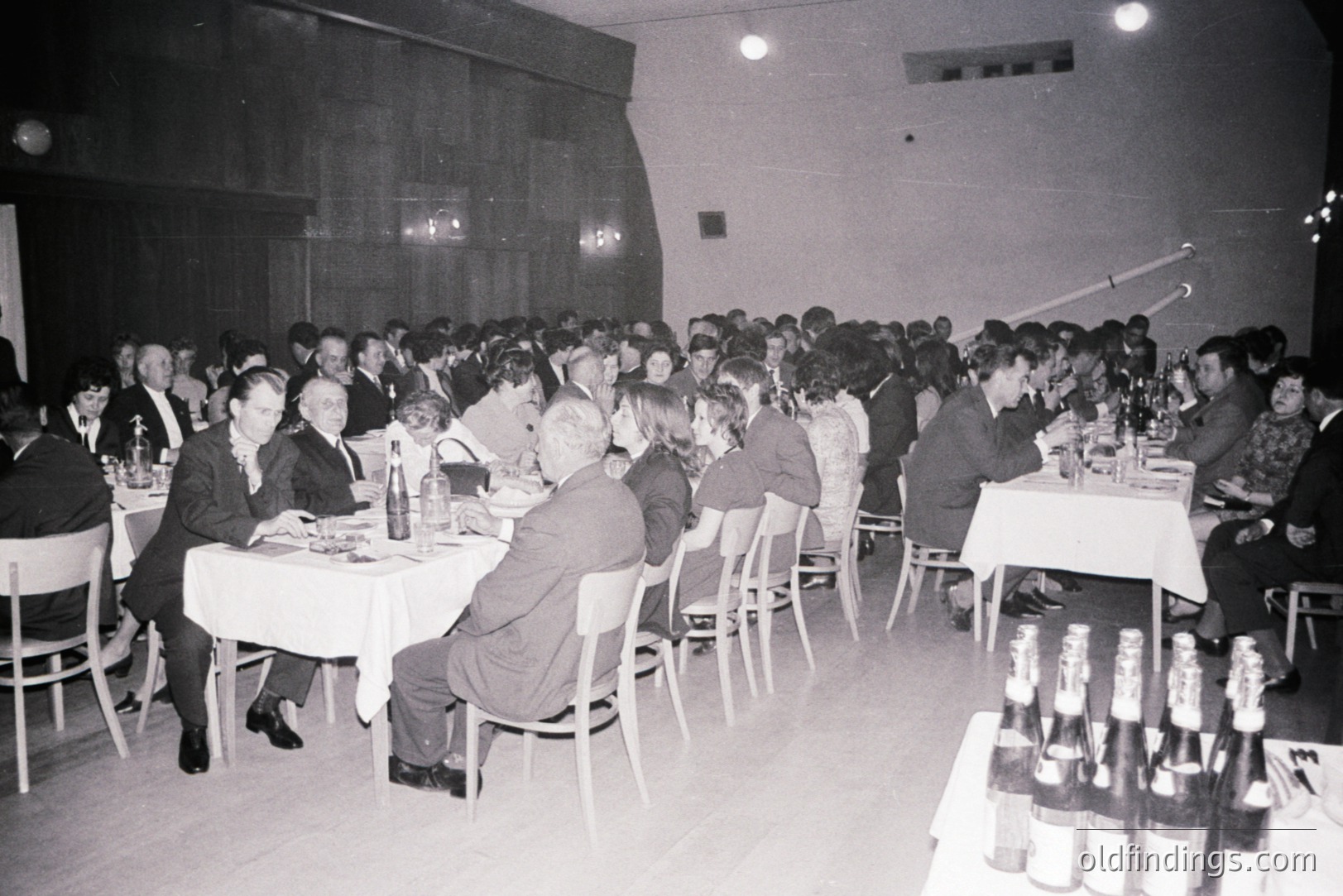 Large group seated at tables covered with white cloths in a hall. Men in suits and ties, some women in dresses. Bottles of wine stacked near tables. Sparse decor, likely a formal gathering, possibly a conference or banquet. Possibly mid-1960s. Venue’s architecture features a curved ceiling.
