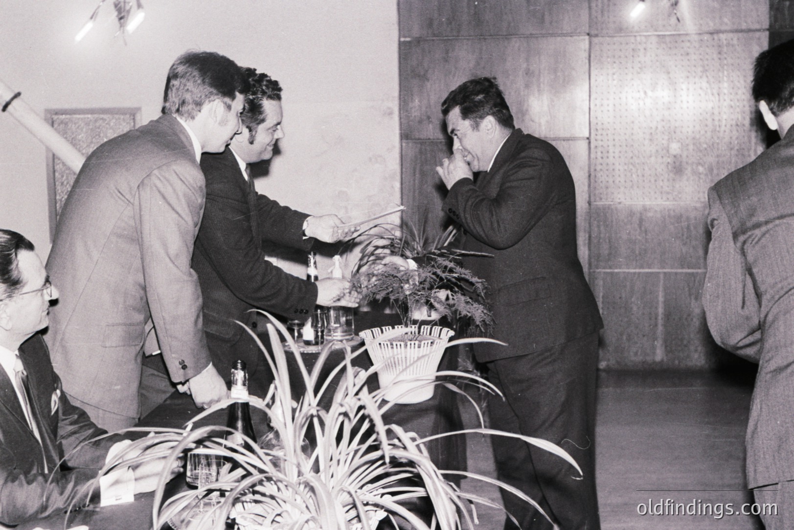 Formal gathering, circa 1960s. Three men in suits observe another man seated, appearing to present a small trophy or award. Indoor setting with textured wall paneling and decorative plants. Likely a corporate event or professional association meeting.