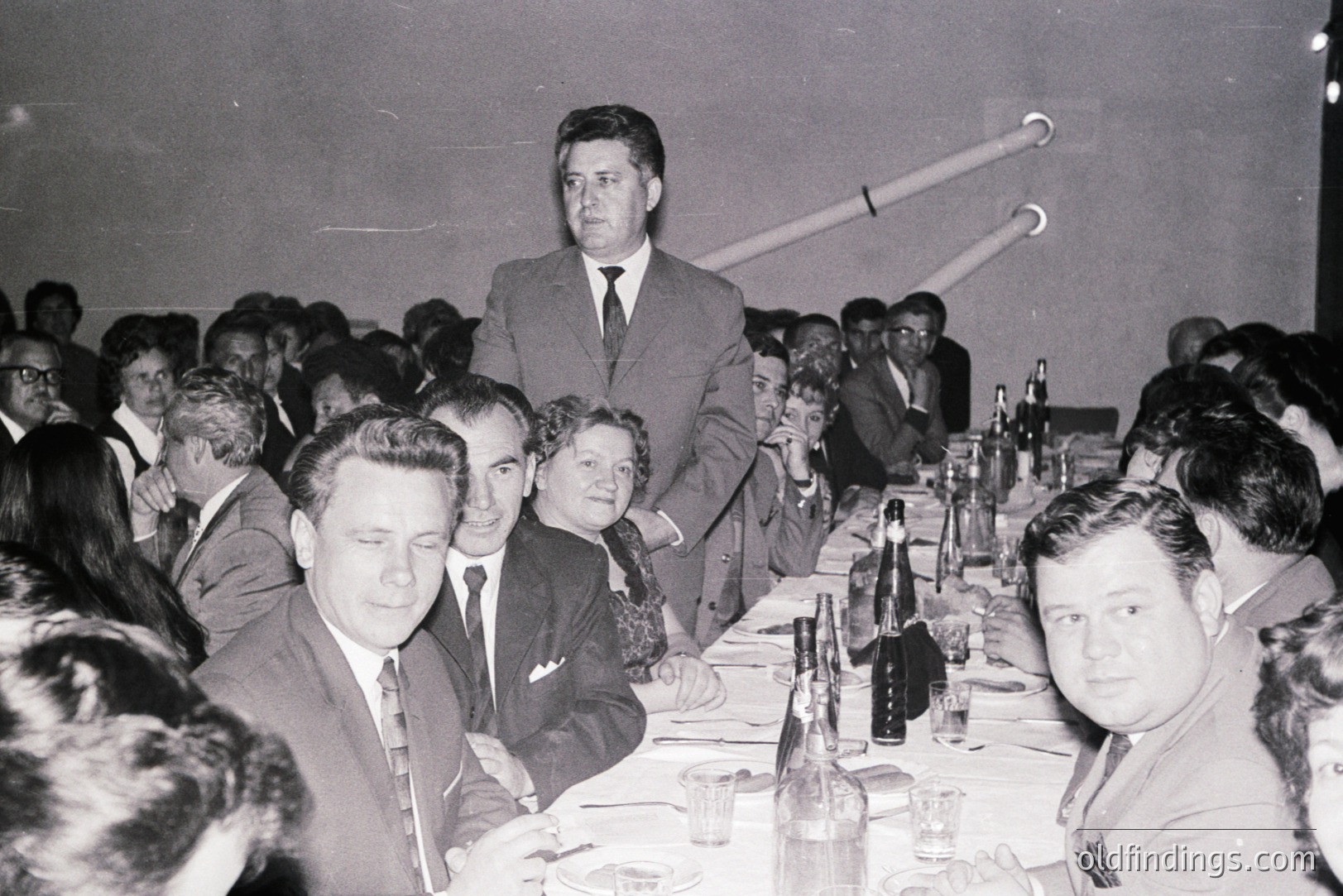 A formal dinner or banquet scene, likely 1960s. Guests seated at tables adorned with dark bottles & glassware. Men in suits, women in formal dresses. A gentleman stands, appearing to address the room. Black and white photography, possibly a documentary or event record.