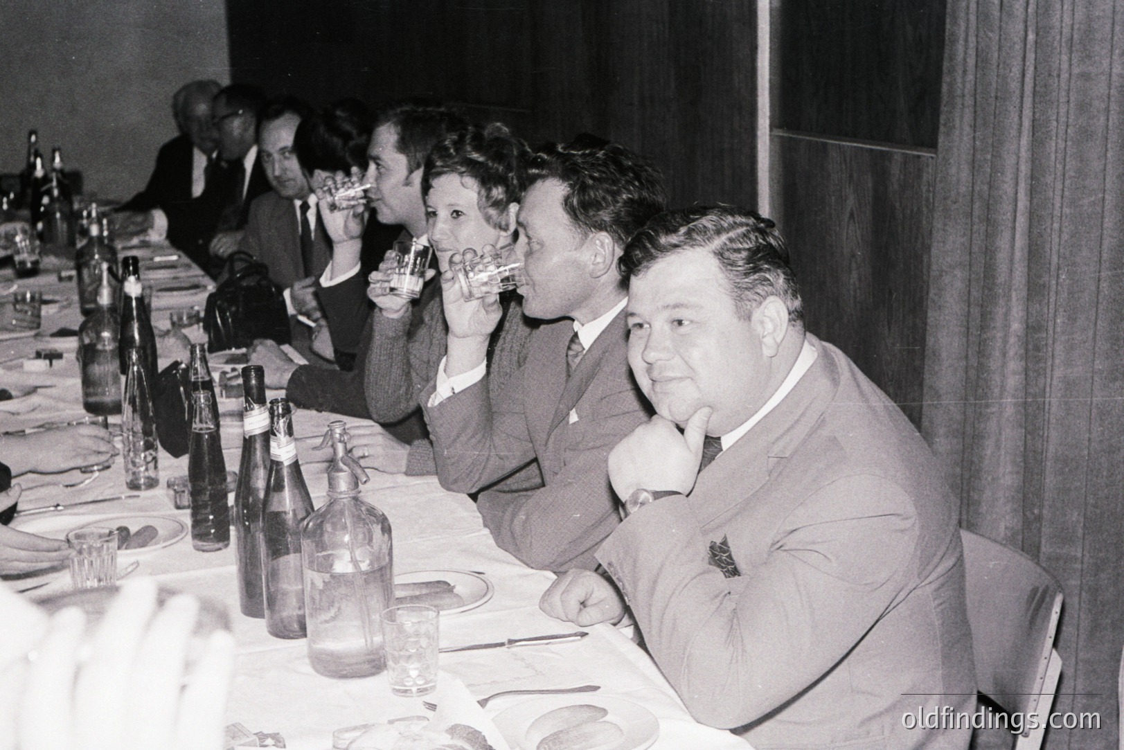 Group scene at a formal dinner. Several men and women in suits and dresses raise glasses in a toast. A long table covered in a white tablecloth is visible. Dark background suggests an event hall or banquet room. Likely 1960s, potentially a business or celebratory gathering.