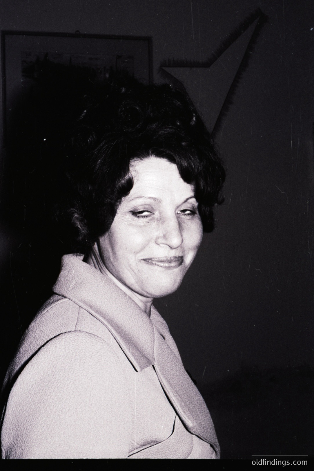 A woman with a warm smile and elaborately styled hair is captured in a portrait. She wears a light-colored, collared jacket. The image, likely taken in the 1960s or 70s, has a grainy quality characteristic of film photography from that era. Background features a blurred architectural detail.