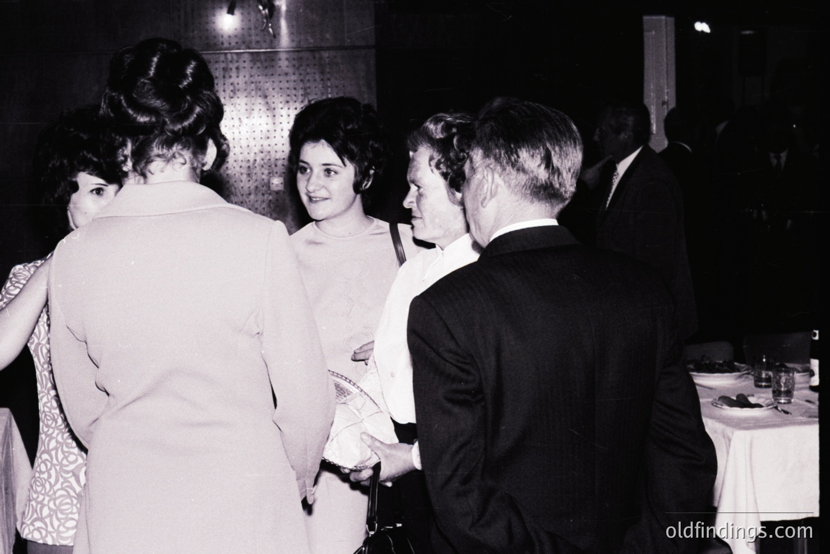 Group portrait captures a formal gathering, possibly a banquet or event. Four figures are prominently displayed: three women in evening wear and a man in a dark suit. The setting appears to be a dining hall with tables set for a meal. Architectural detail: corrugated wall. Likely 1960s.
