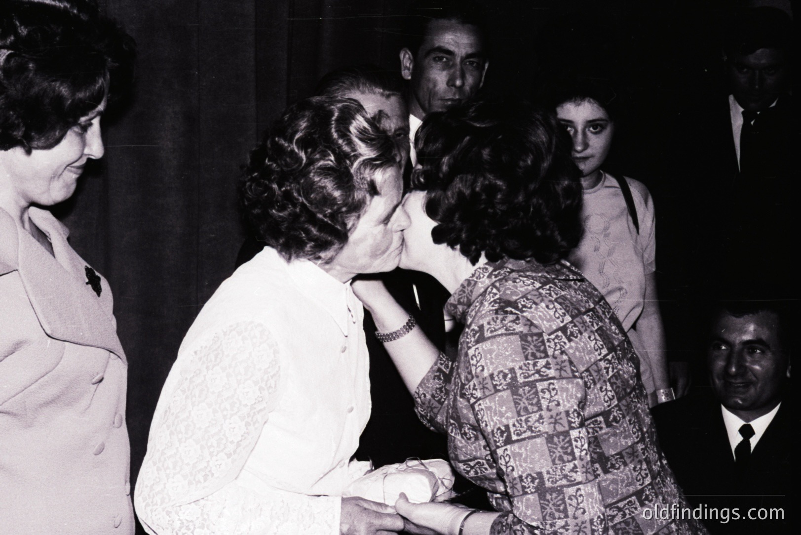 A formal, intimate moment captured: a woman in a white, lace-trimmed dress receives a kiss on the cheek from another woman with patterned clothing. Several onlookers are present, slightly blurred, suggesting a public or semi-formal setting. Likely 1960s-1970s.