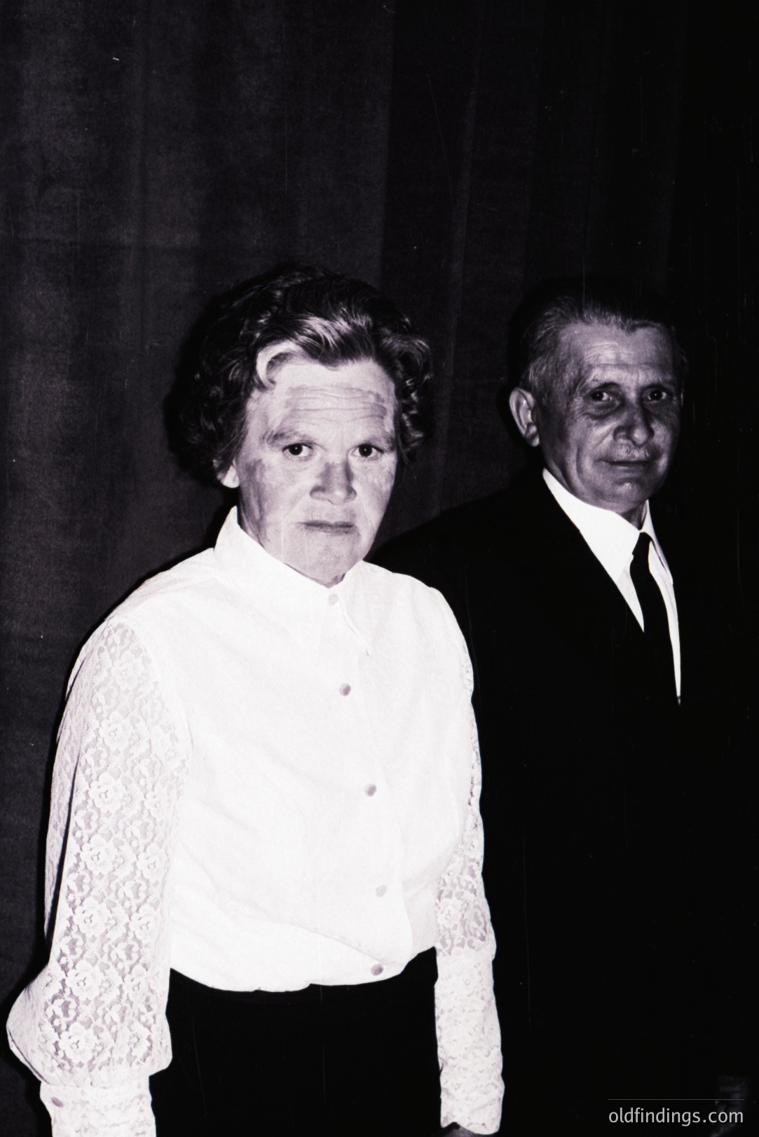 A striking black and white portrait showing a mature woman and gentleman. The woman wears a lace-collared blouse and dark skirt, standing prominently. The man, in a suit and tie, stands slightly behind her. Likely a formal event setting. Evokes a mid-20th century aesthetic.