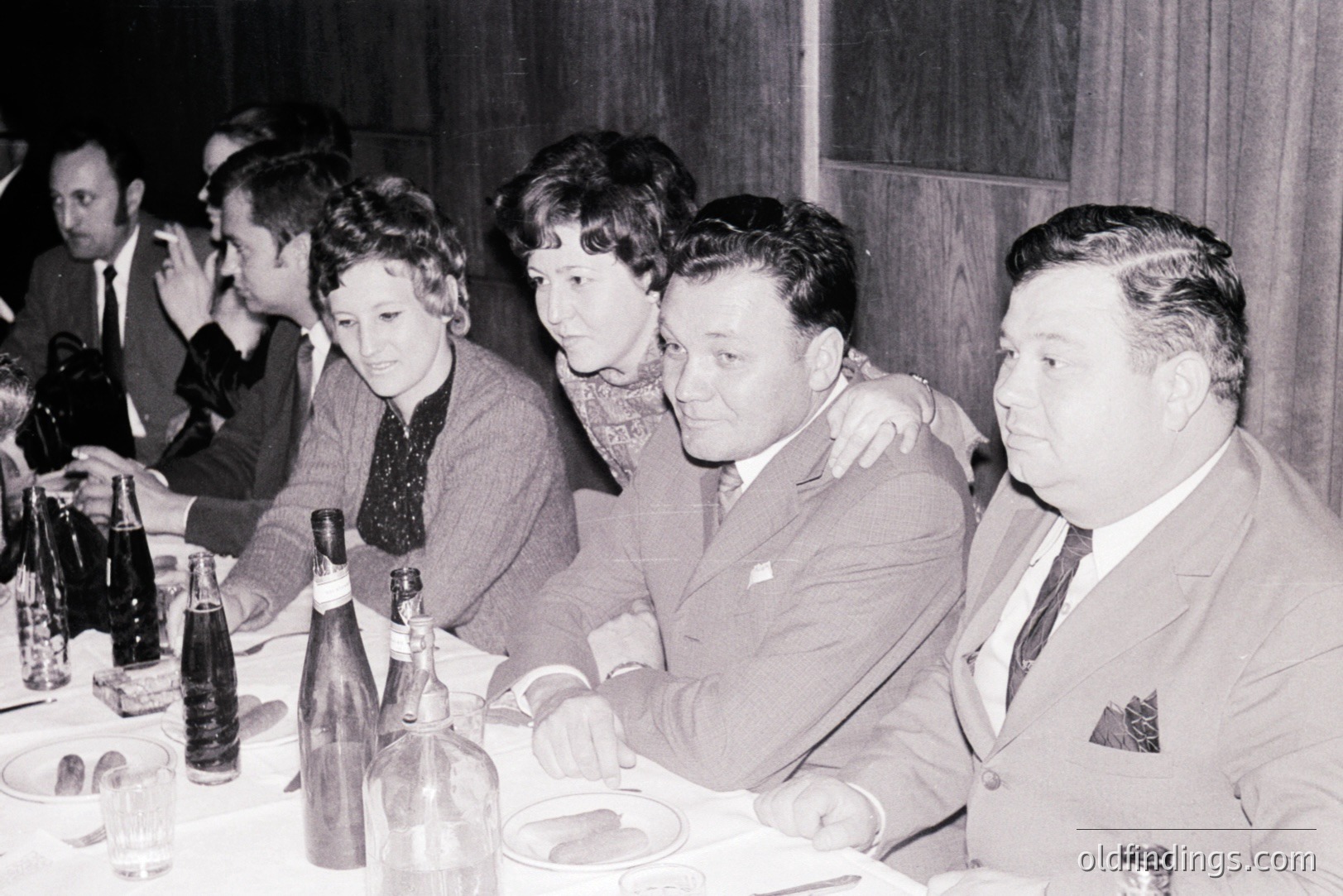 A candid group portrait at a formal dinner. Four men and two women are seated at a long table, enjoying a meal with drinks and décor visible. The men are in suits, while the women wear dresses or blouses. Likely a 1960s or 70s event.