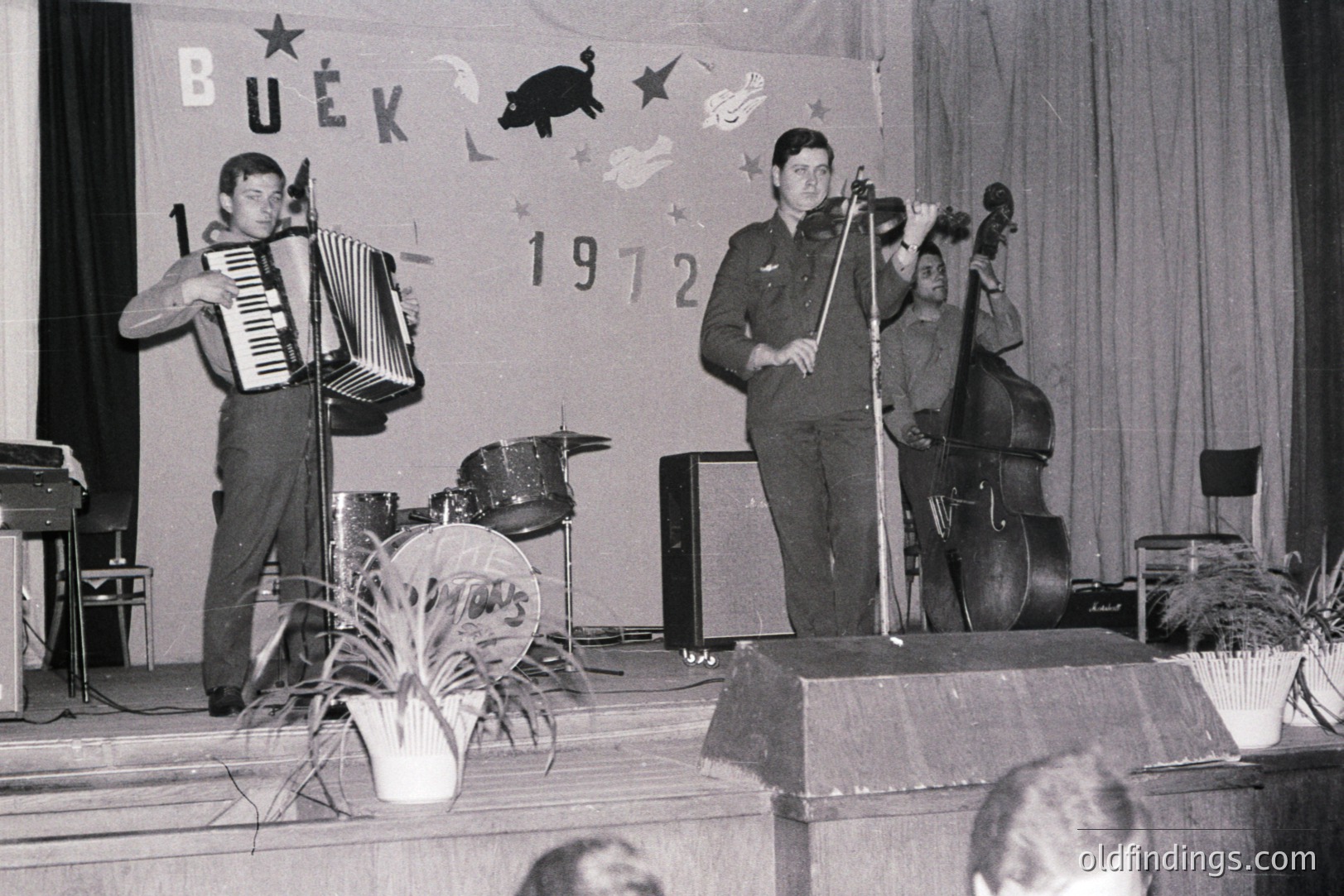 Three musicians perform onstage, featuring an accordionist, bassist, and drummer. A backdrop displays "BUEK" and "1972." Stage decor includes potted plants and a vintage amplifier. Likely a student performance or local cultural event.