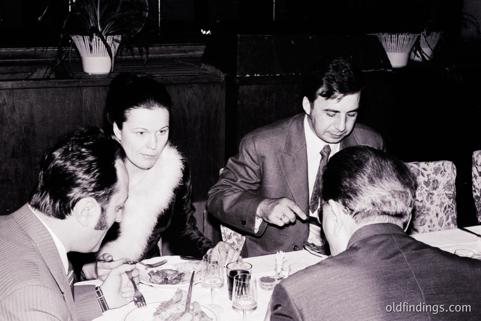 Four formally dressed individuals seated around a table, likely dining in a restaurant. A woman with a fur collar is central, while men in suits engage in conversation. Appears to be a candid, mid-century capture. Possible social or business gathering.