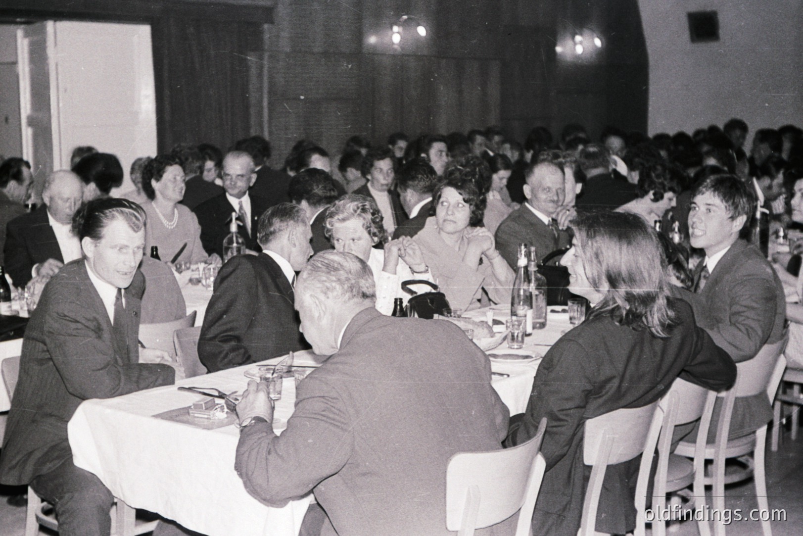 A formal gathering, likely a banquet or business event. Numerous formally dressed attendees sit at tables covered with white tablecloths. Men wear suits; women, modest dresses. The setting appears to be a large hall with institutional lighting. Appears to be 1960s-1970s.
