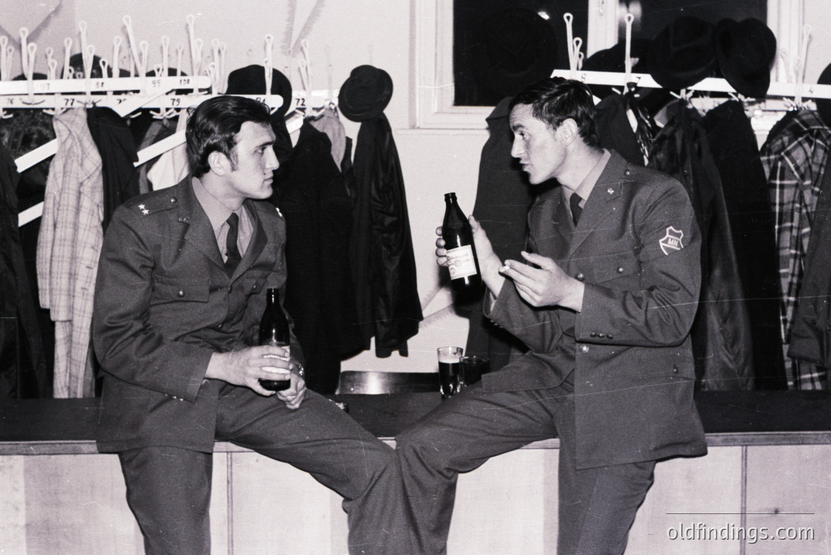 Two uniformed men, likely military personnel, are seated and holding bottles, perhaps of beer or wine. Background shows a storage area with racks of clothing and hats. Appears to be an informal moment, possibly off-duty. Likely 1960s-1970s, origin unknown. Offers visual reference for military uniforms & mid-century leisure.