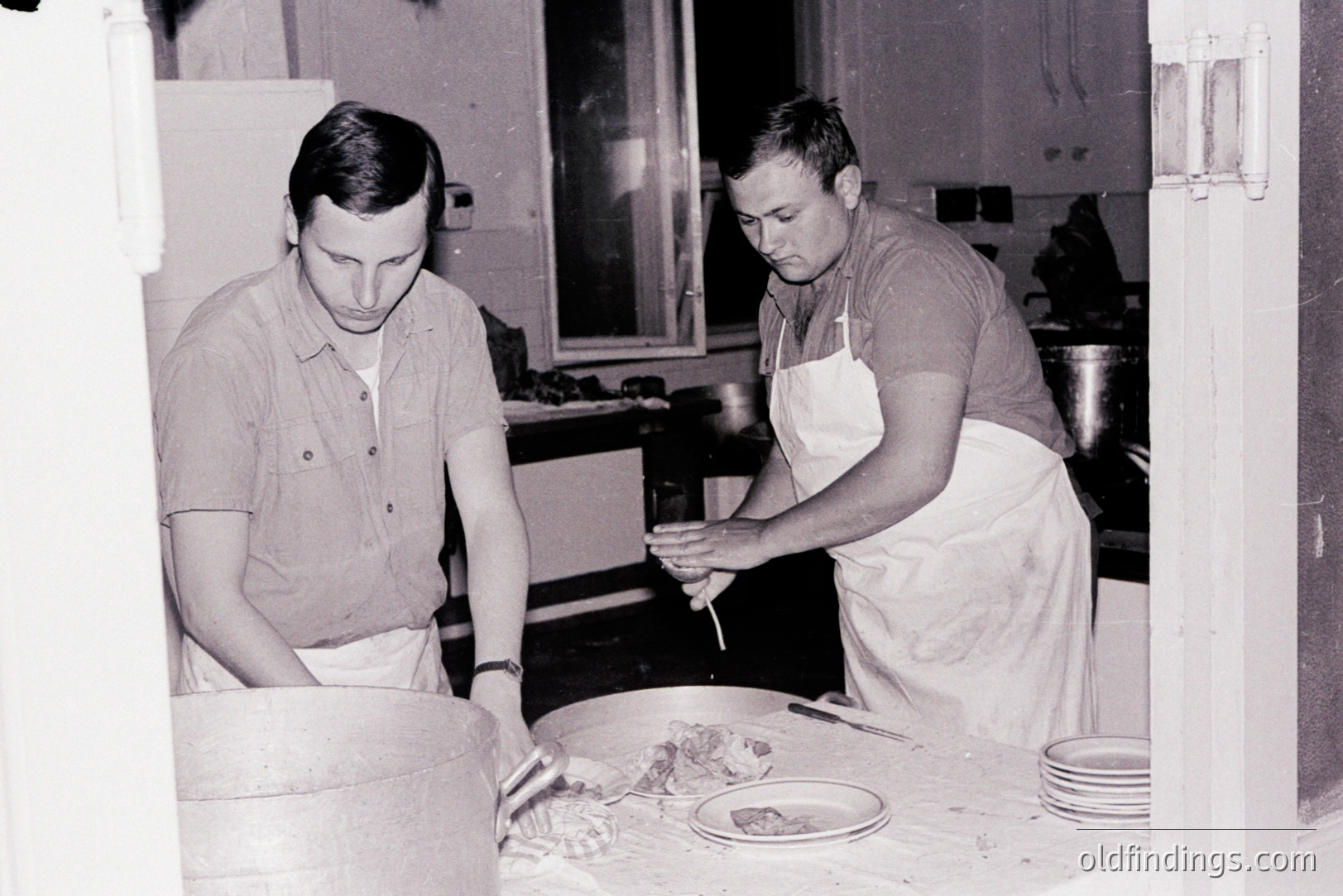 Two men in short-sleeved shirts and aprons are shown in a workspace, likely preparing food. One kneads dough in a large wooden container; the other trims or prepares a meat dish on a round wooden board. Simple, functional environment suggests a farm or small business setting. Appears to be mid-20th century.
