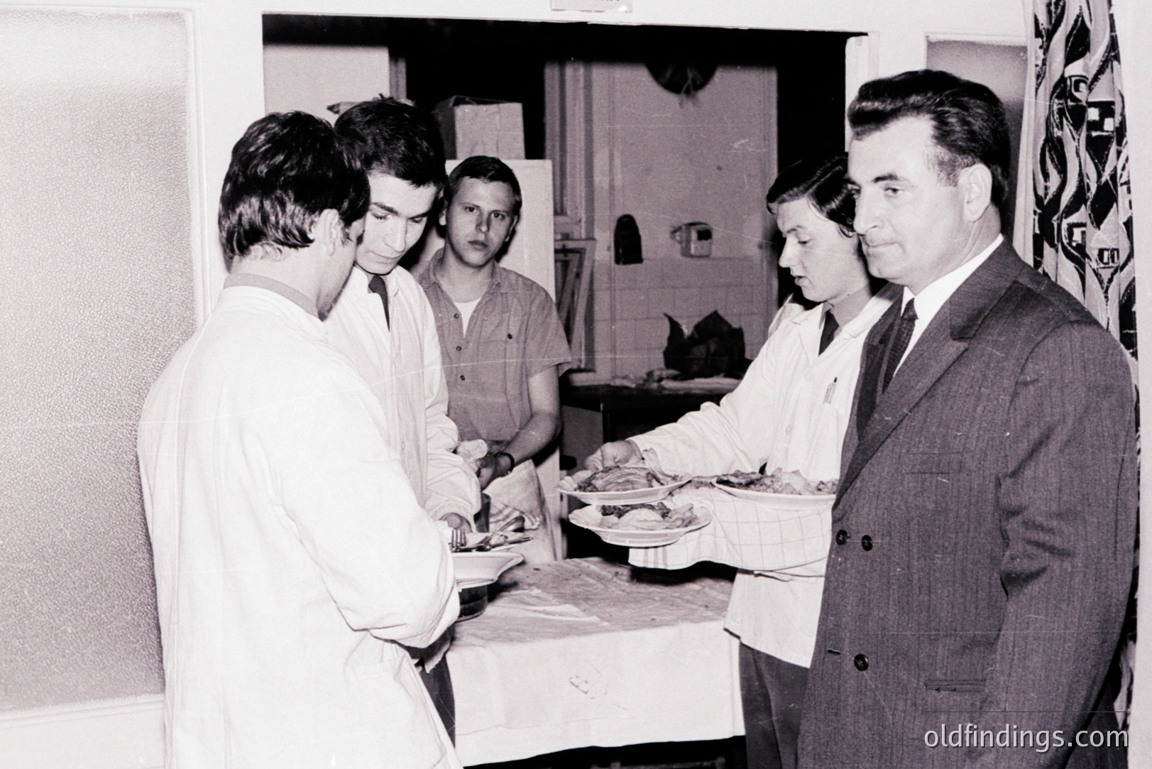 A formal gathering; a man in a suit observes two people in lab coats presenting food on trays in a tiled kitchen. Possibly a medical or culinary demonstration, circa 1960s-70s. The patterned wallpaper & decor suggest a domestic interior. Strong potential for historical research & design inspiration.
