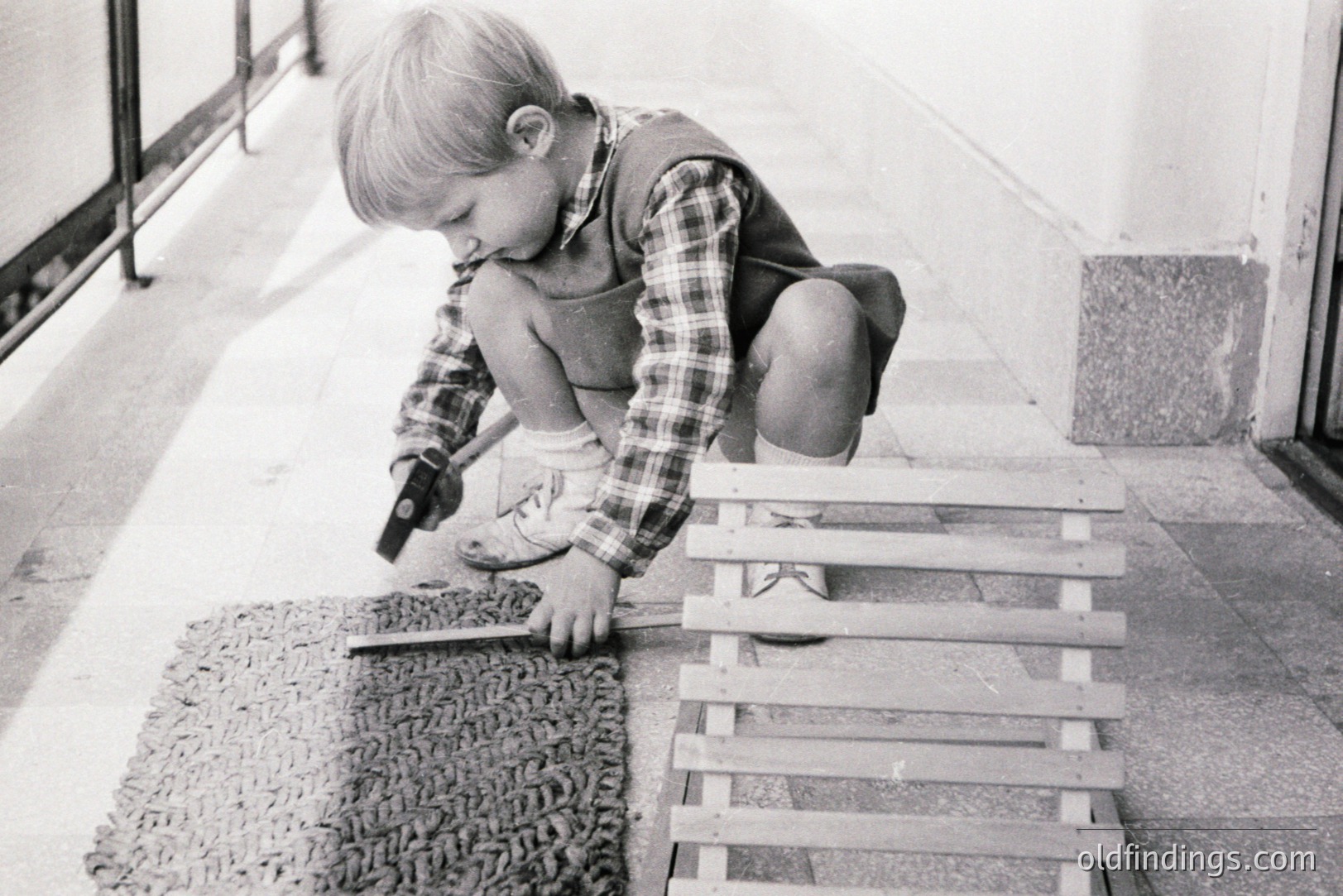 A young boy, crouched on a textured mat, appears engrossed in using a small hammer and tool on a wooden structure. He's wearing checked overalls and simple shoes, likely 1960s attire. The setting appears to be an outdoor patio or terrace. Captures childhood curiosity & early DIY.