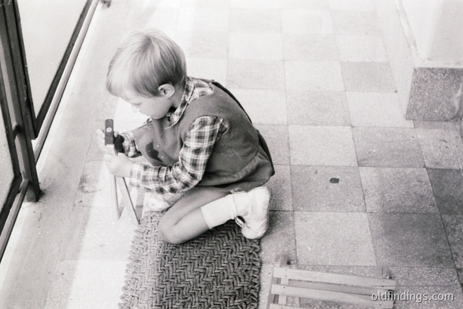 A young boy, possibly 6-8 years old, kneels near a building’s glass entrance, intently focused on a small, boxy camera. He wears a plaid shirt, vest, and knee socks. The tiled floor and textured mat suggest a public building interior. Likely 1960s-1970s. Potential for vintage/nostalgia stock imagery.