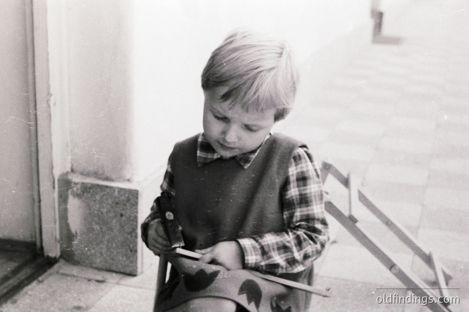 A young boy in a plaid shirt and sweater vest intently examines a piece of wood, perhaps a building block. Seated on a staircase, the image evokes a nostalgic, domestic scene. Likely a snapshot from the 1960s or 70s, its simplicity lends itself to design or historical research.