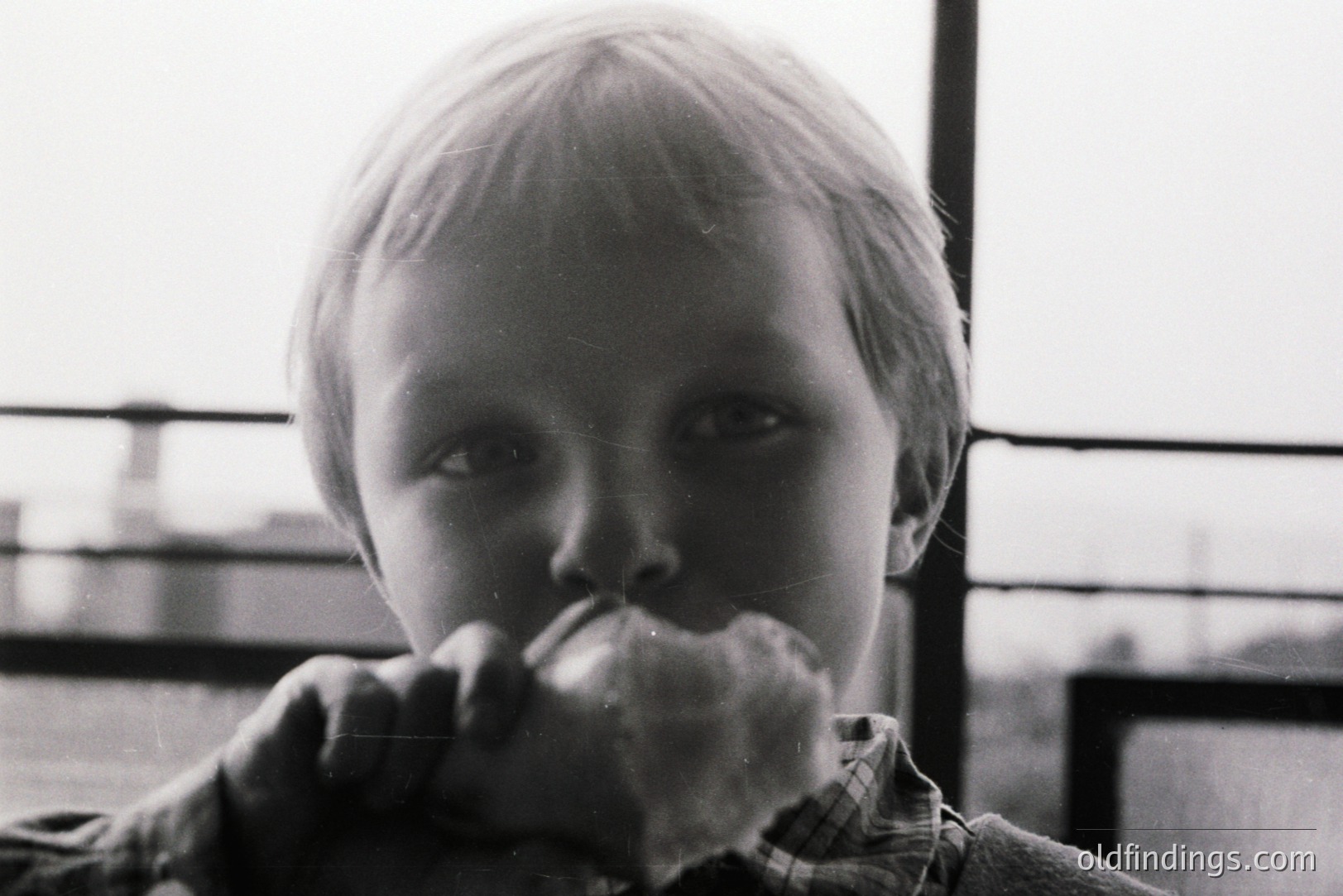Close-up, black and white photo captures a young boy with short blond hair, intently eating what appears to be a large sandwich. View is framed by a metal railing. The plaid shirt adds texture. Likely a casual snapshot, evoking a candid, domestic feel.