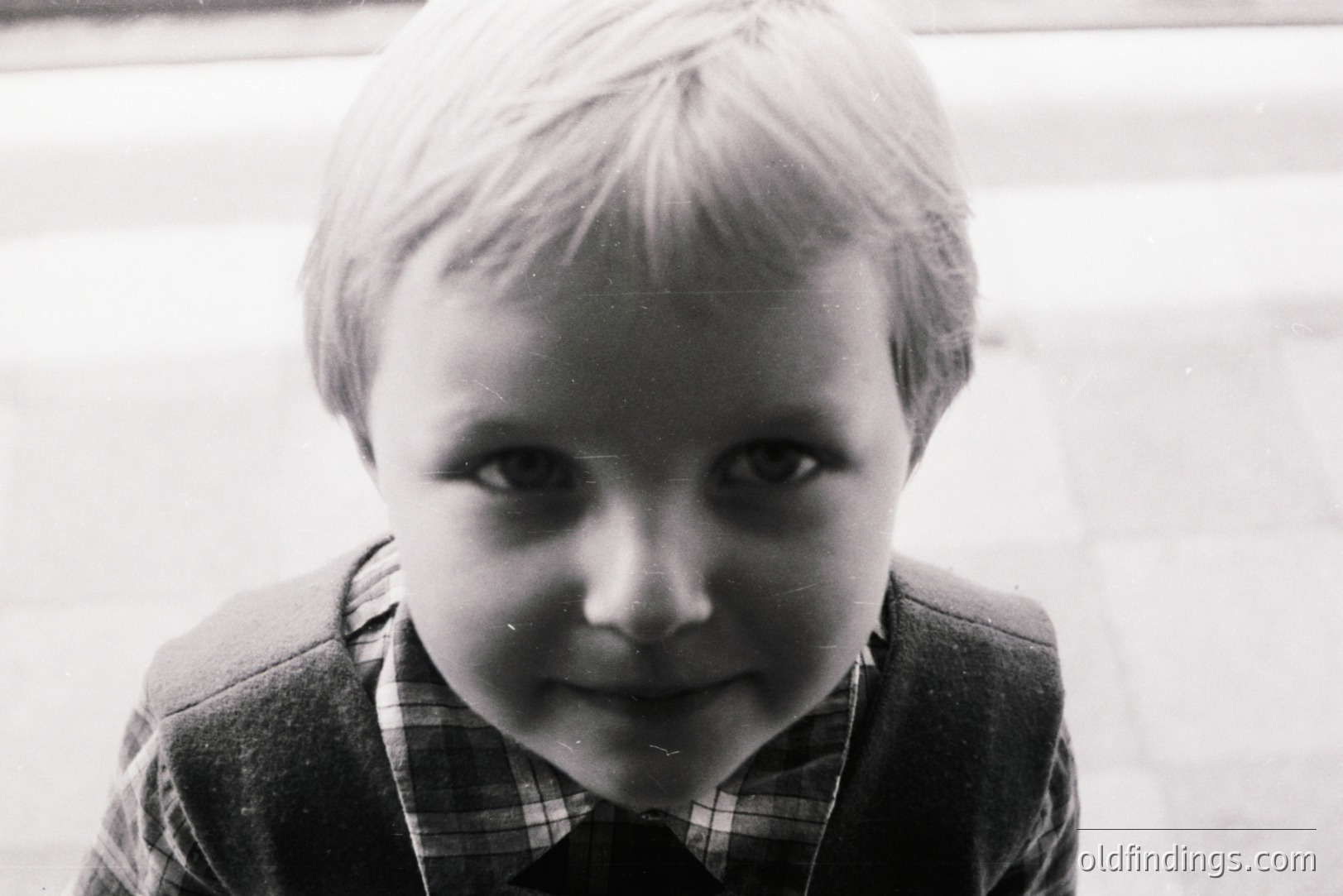 Close-up, candid portrait of a young boy with short, light hair. He wears a plaid shirt and jacket, looking directly at the camera with a slight smile. Grainy texture suggests film photography, likely 1960s-1970s. Evokes a sense of childhood nostalgia. Potential for design or archival use.