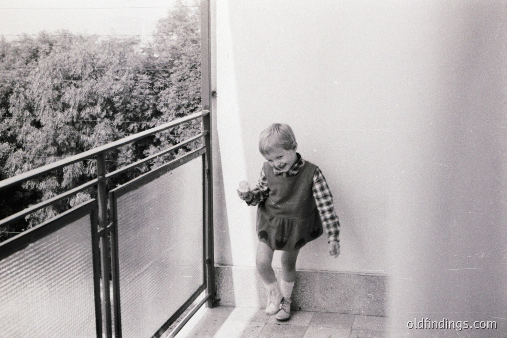A young boy in a checked shirt and bloomers playfully poses on a balcony. The black and white image likely dates to the 1960s or 70s, showing a glimpse of greenery beyond the railing. A simple, functional design aesthetic is evident. Possibly a family vacation photo.