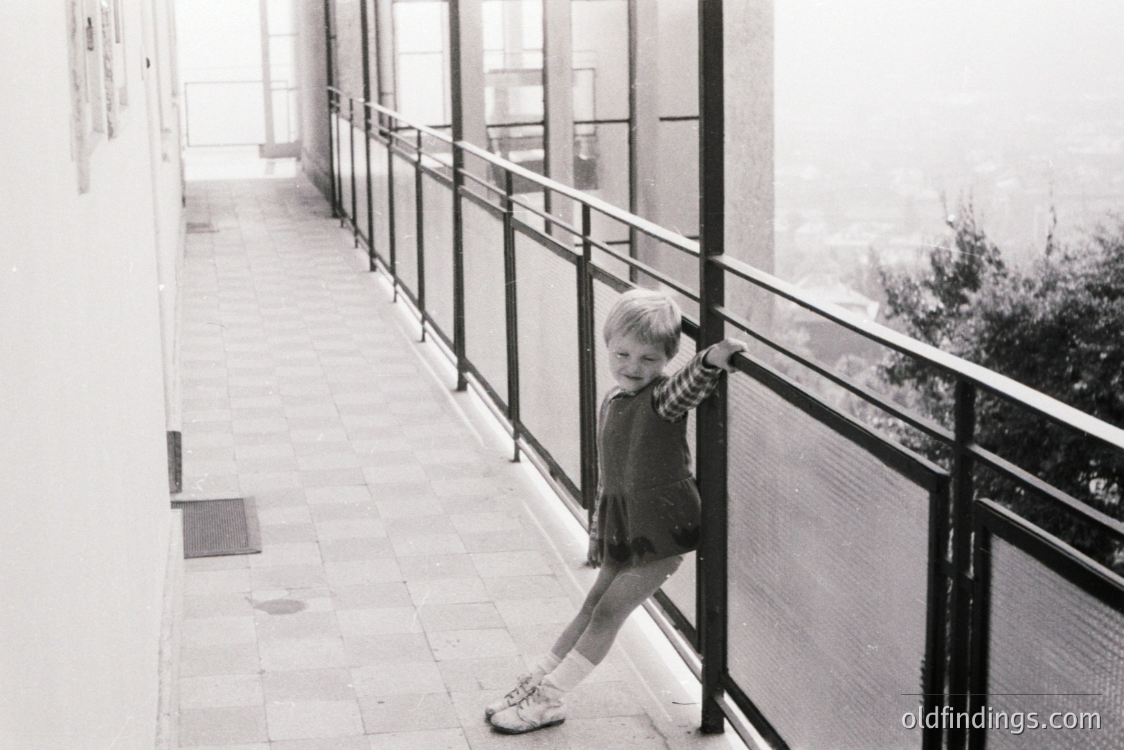 A young child stands playfully on a balcony, posed with one arm on the railing. The tiled floor and dark metal railing suggest mid-century modern architecture. A blurred landscape stretches out beyond, hinting at coastal or mountainous terrain. Likely 1960s, possibly European. Stock photo reference for architectural or childhood themes.