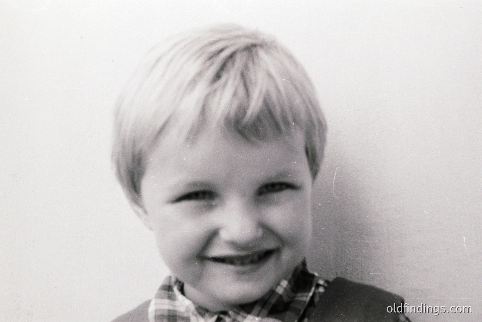 Close-up portrait of a young boy with short, blond hair and a cheerful grin. He's wearing a plaid shirt and a jacket. Plain background suggests a studio or simple home setting. Likely a candid snapshot from the 1960s or 1970s. Good for family archives & nostalgic design.