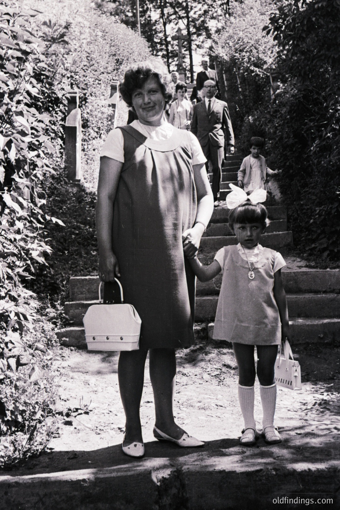 A woman and young girl stand on stone steps, holding hands, alongside a group ascending in the background. The woman wears a distinctive, A-line dress and carries a white, boxy handbag. The girl wears a matching dress, knee-high socks, and a large bow. Likely 1960s, possibly Eastern European due to the clothing styles. A candid family moment, potentially valuable for fashion or historical research.