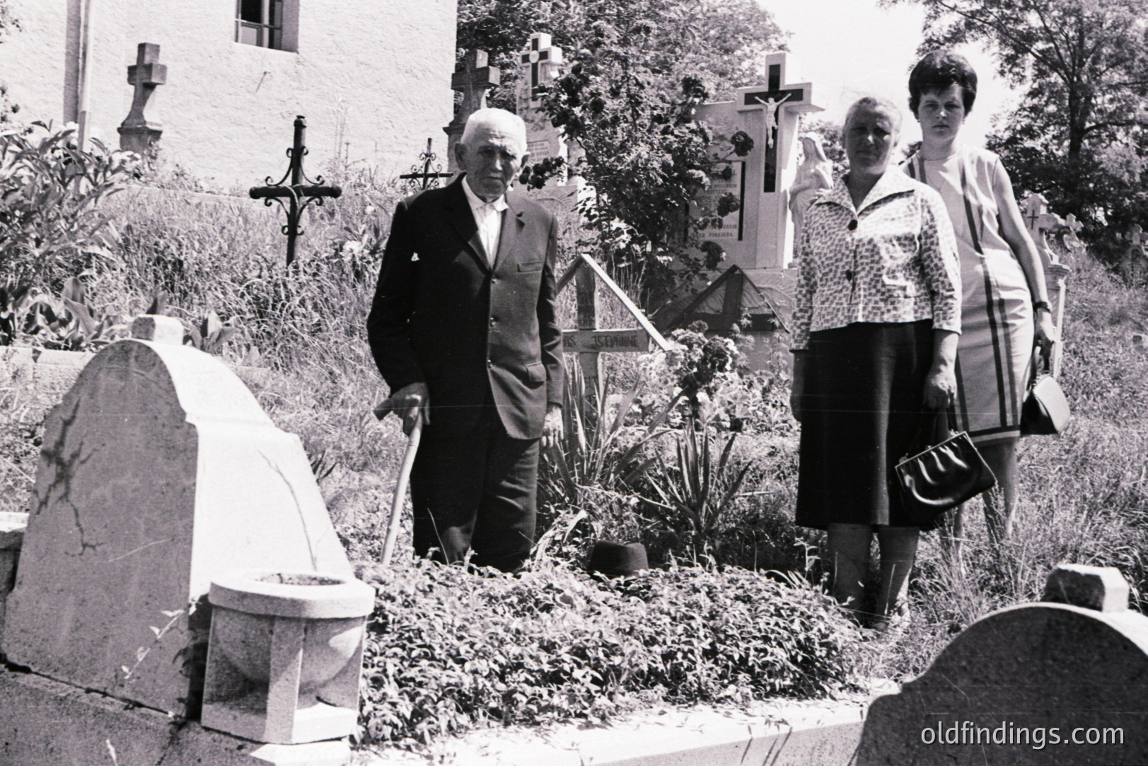 Monochrome image of an elderly man in a suit and two women in 1970s attire walking through a graveyard with ornate headstones and a stone building in background. Likely Eastern European, possibly Bulgaria, judging by the architectural style. A poignant glimpse of a family visit.