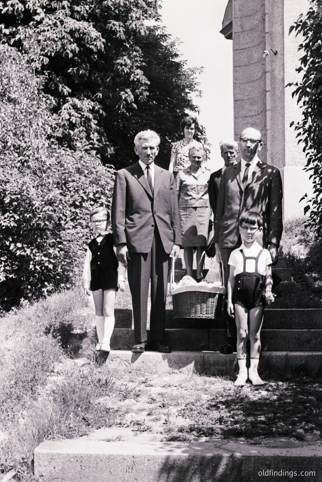 Formal portrait, likely 1960s. Family ascends stone steps; father and son in suits, mother & daughter in patterned dress & pinafore. Woman carries a wicker basket. Architectural elements suggest a European estate. Crisp, slightly grainy monochrome. Strong visual potential.