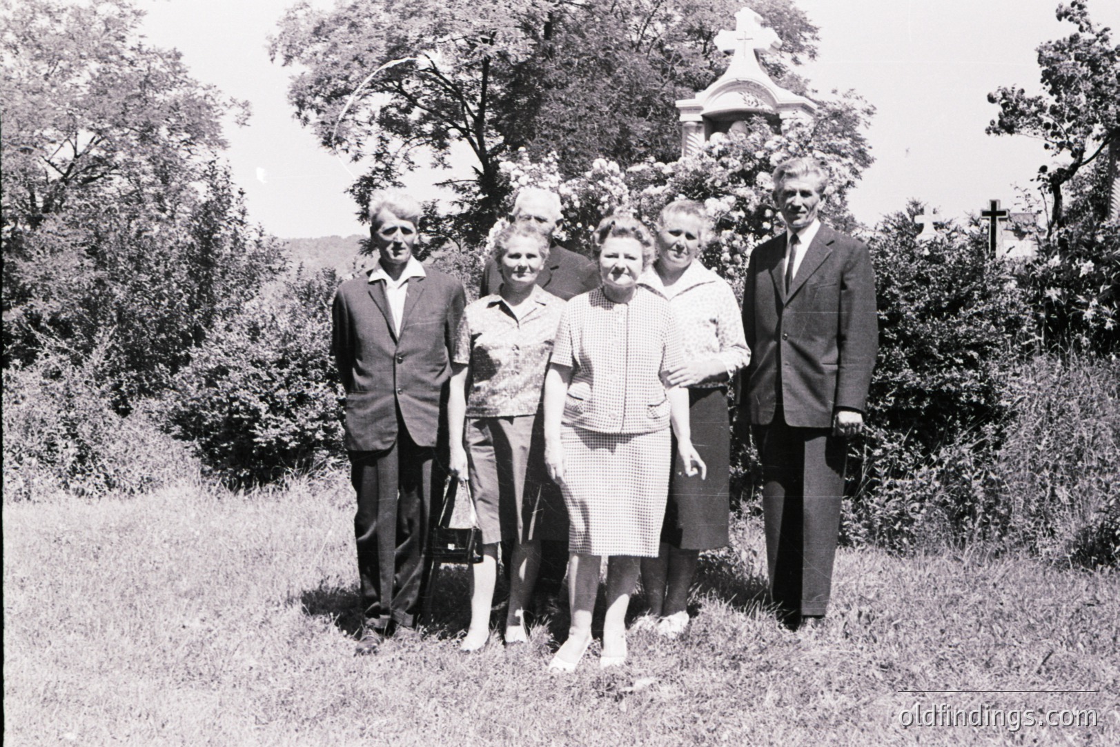 A formal portrait of five individuals, likely a family, posed in a grassy area with a small, ornate chapel visible in the background. Men wear dark suits & ties; women in dresses/skirts. Appears to be a mid-century snapshot, potentially 1950s-1960s. Possible Eastern European setting.
