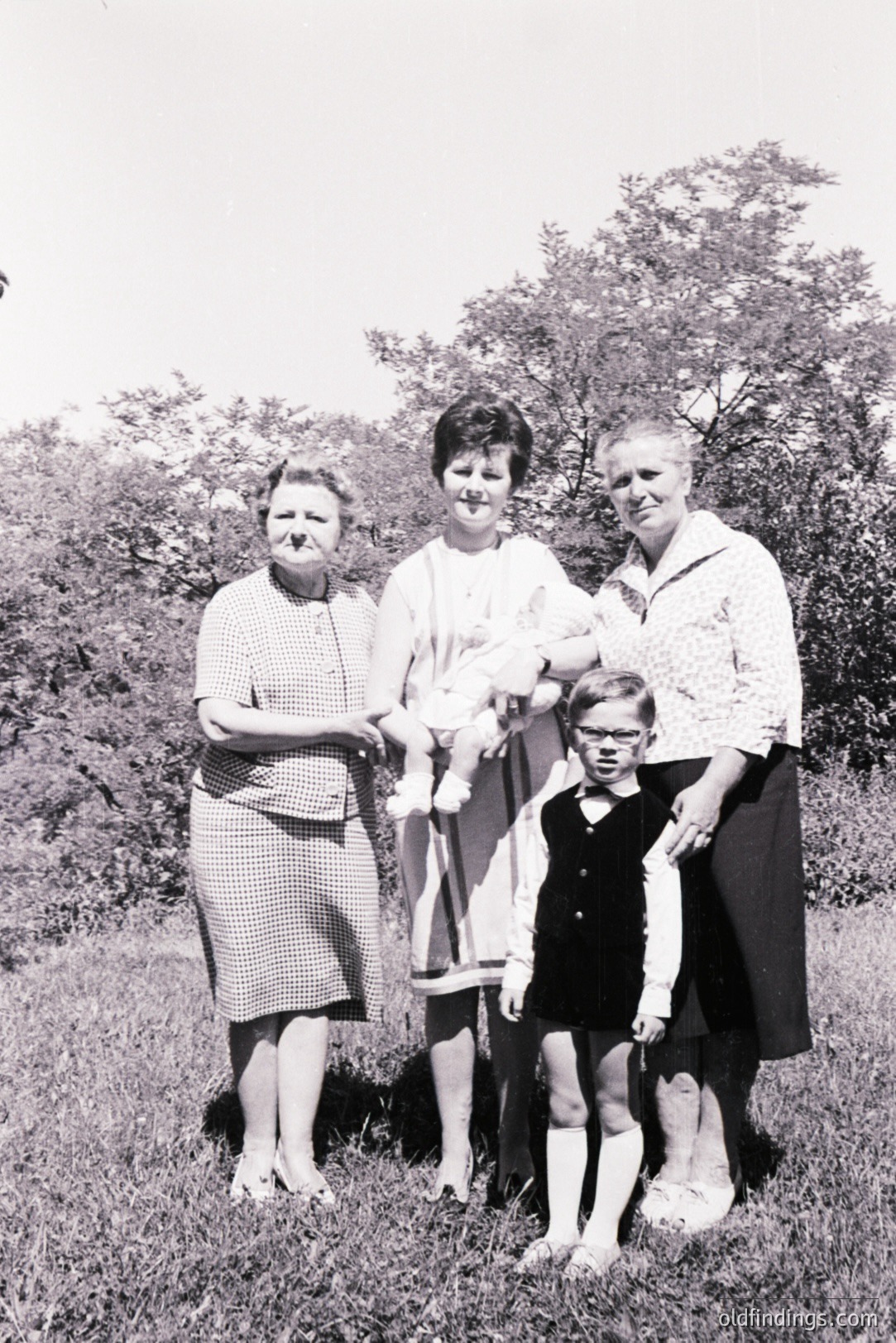 Family portrait, likely 1960s. Three women and a young boy stand in a grassy field with trees in the background. The women wear vintage dresses/skirts & stockings. Boy wears glasses and short shorts with knee-high socks. Likely a posed, informal snapshot.