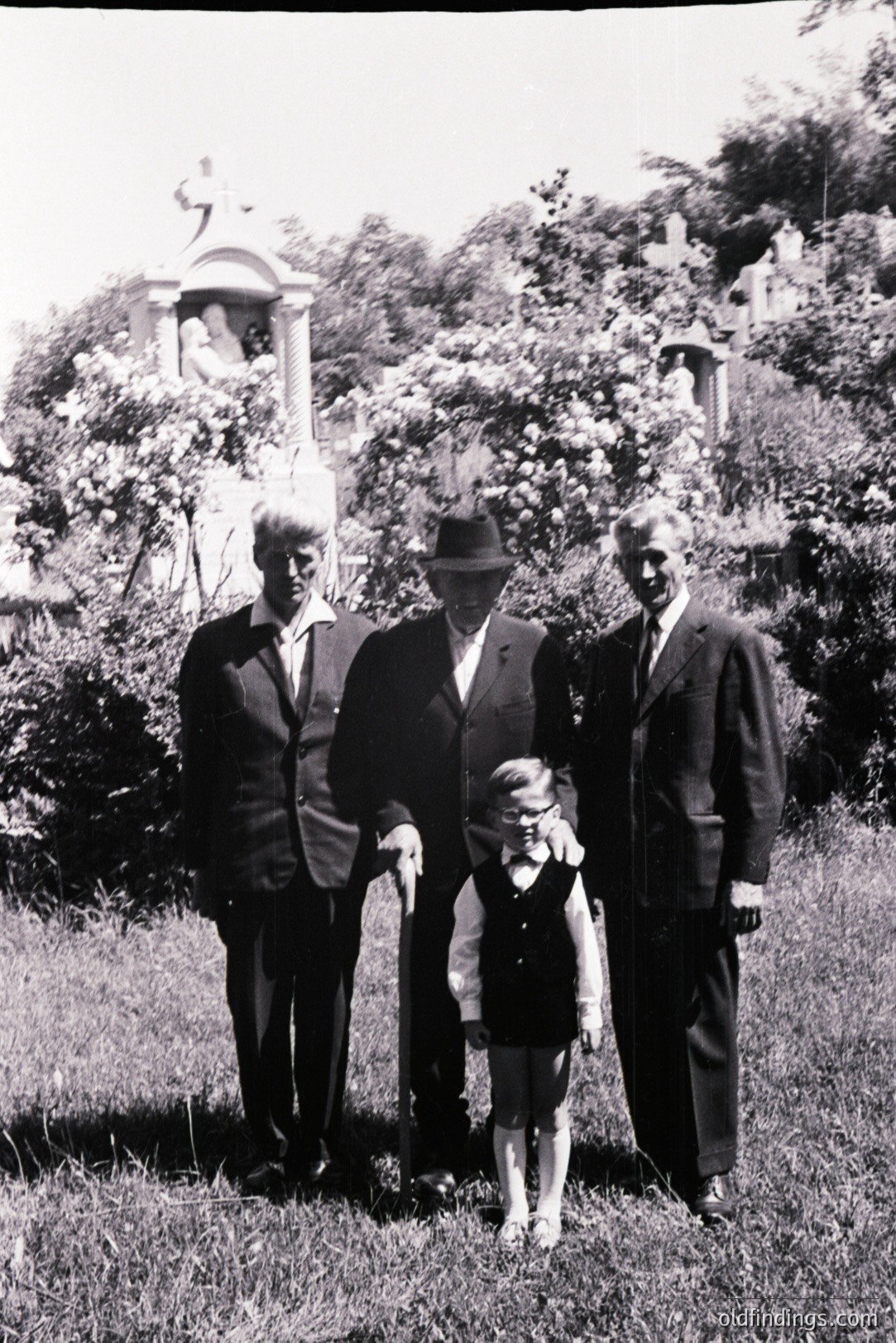 Formal portrait of three men and a young girl in a cemetery setting. Architectural details include a stone monument, likely a mausoleum, visible through flowering shrubbery. Subjects are formally dressed in suits and a hat; girl wears a dress with bow. Likely 1950s-1970s. A valuable historical family image.