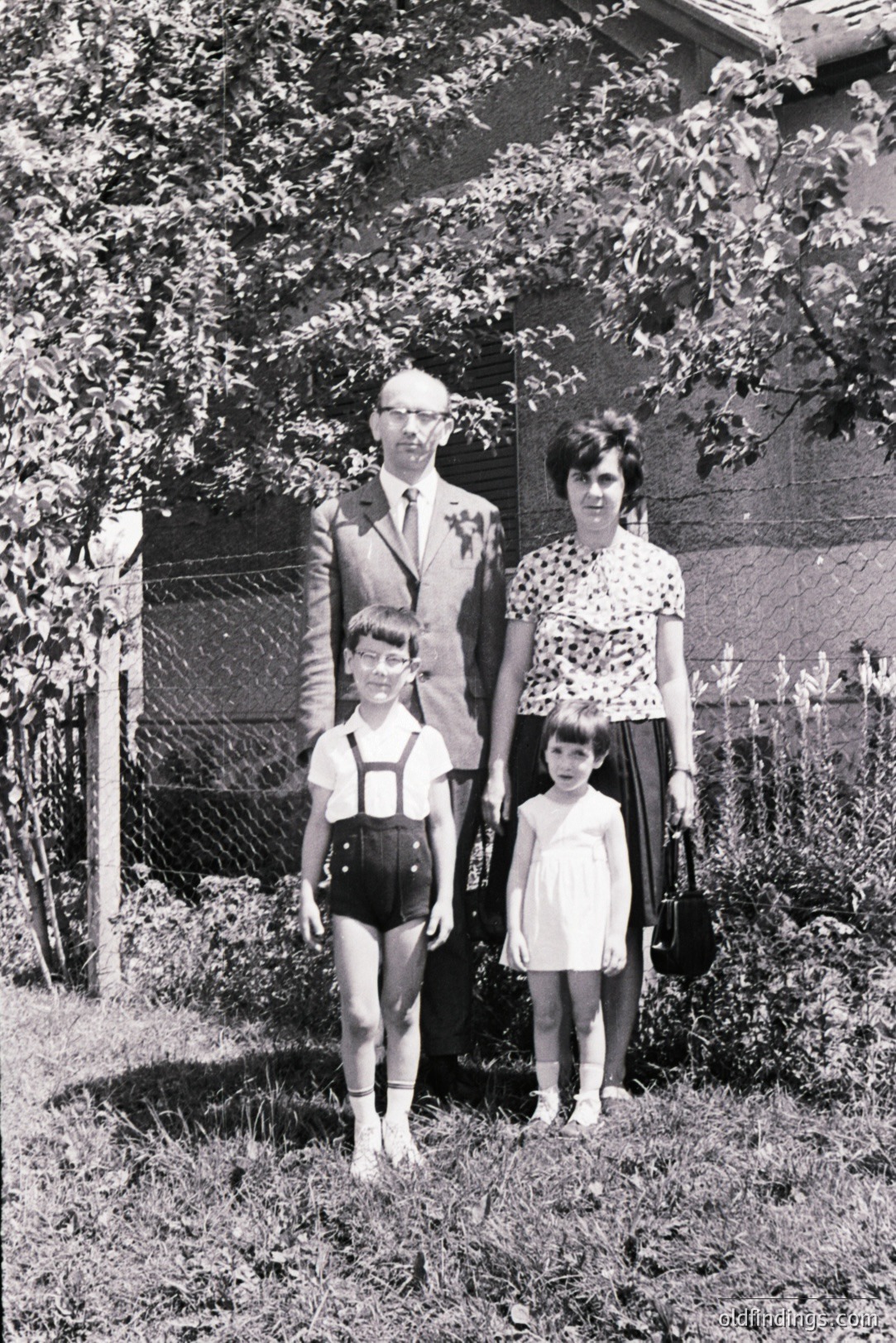 Family portrait, c. 1960s. A father in a suit, mother in a patterned dress, and two young children stand in front of a modest home. The boy wears overalls, the girl a simple dress, both barefoot. Likely a middle-class family snapshot. Offers nostalgic value.