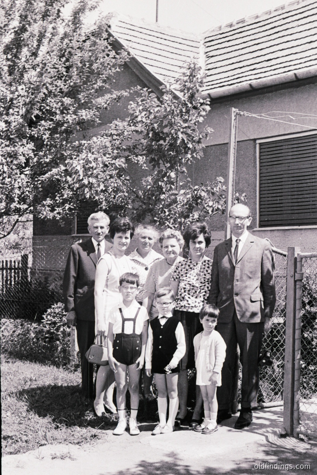 Family portrait, circa 1960s, shows three generations posed in front of a modest, tiled-roof home. Men wear suits & ties, women dresses, children in short pants & overalls. A natural, overgrown garden backdrop suggests a rural or suburban setting. Evokes nostalgic family moments.